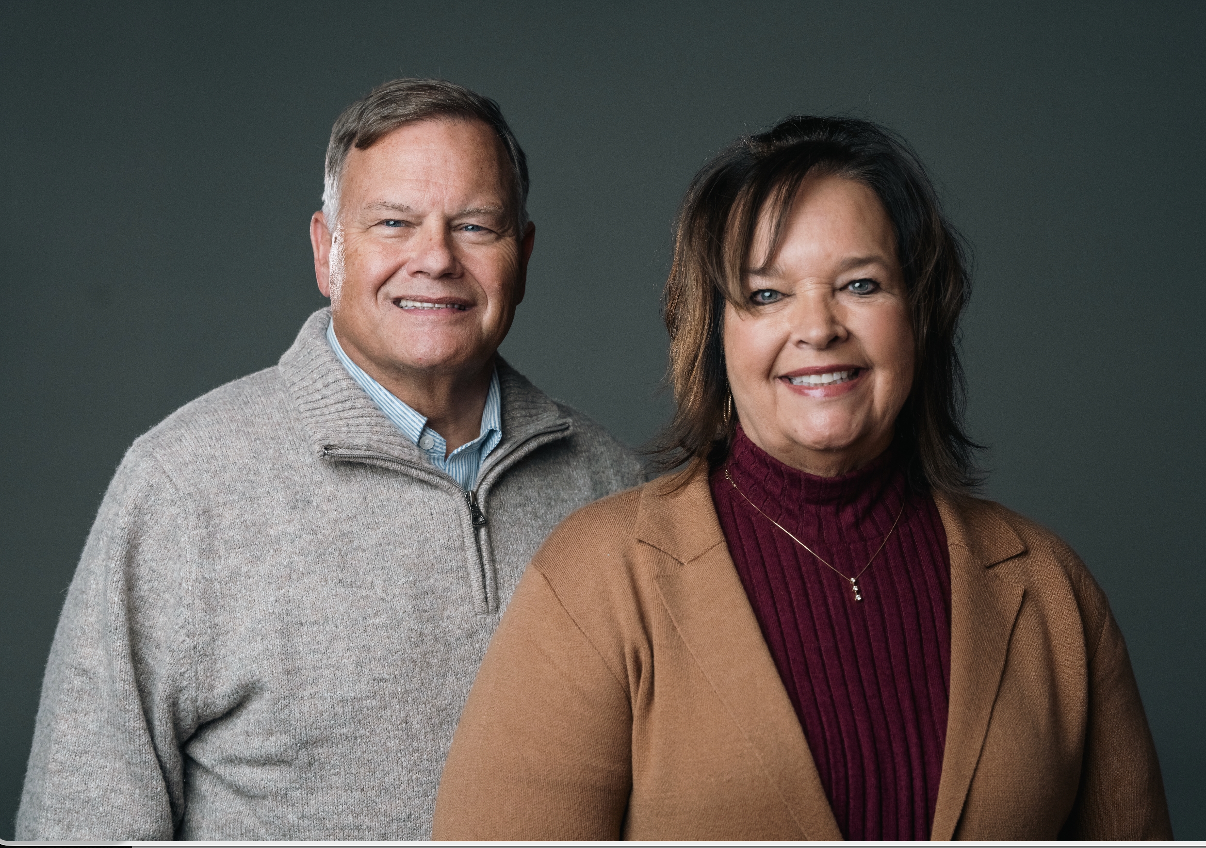 A middle-aged man and woman standing together, smiling against a plain beige background. The man is wearing a light gray button-up shirt, and the woman is wearing a cream-colored sweater.