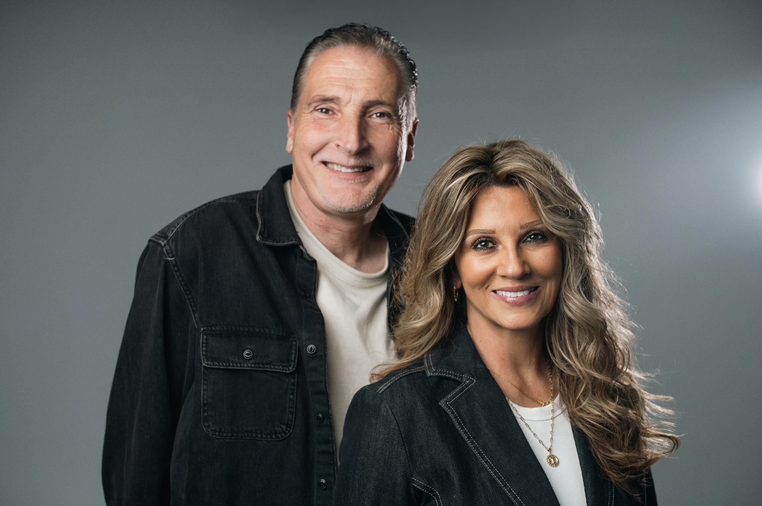 A man and woman standing together, both smiling, in a studio with a plain beige background. The man has gray hair and is wearing a denim jacket with a shearling collar, and the woman has long, wavy blond hair and is wearing a tan blouse with lacework.