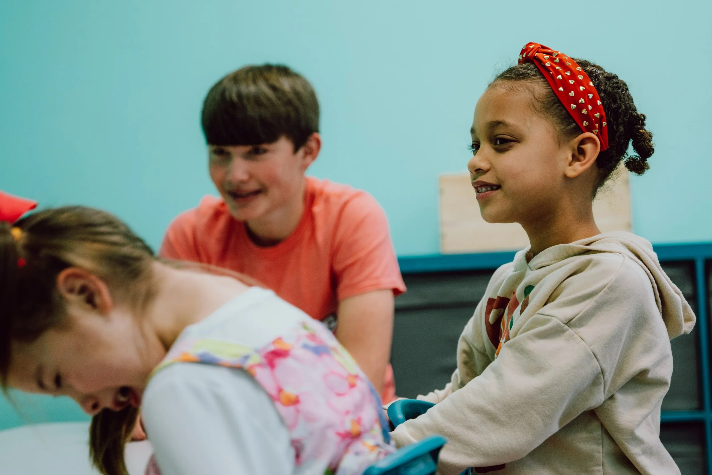 Children laughing and playing together indoors, with a girl wearing a red headband and a brother in a coral T-shirt.