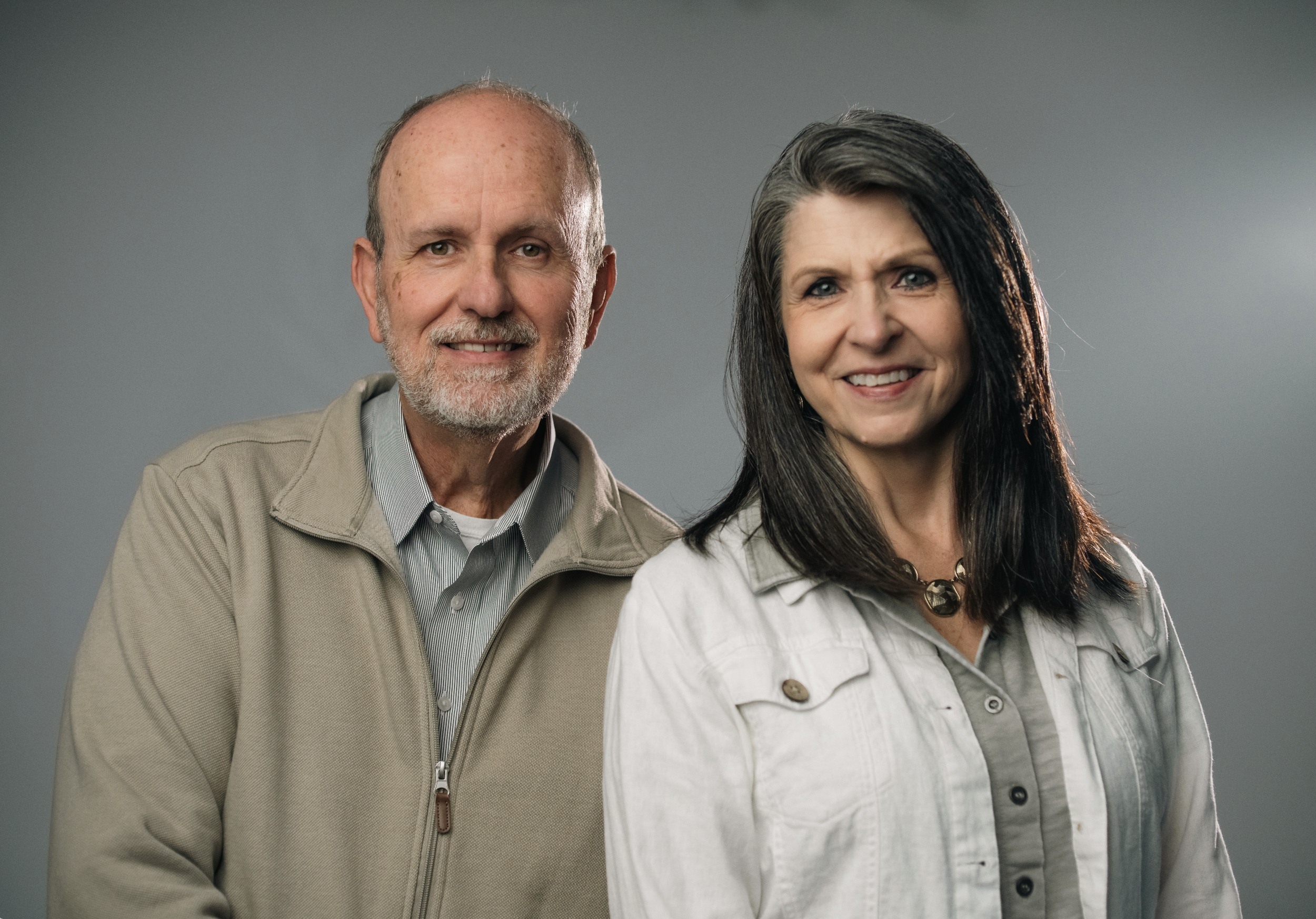 A middle-aged man with glasses and a beard standing close to a middle-aged woman with shoulder-length dark hair, both smiling, against a beige background.