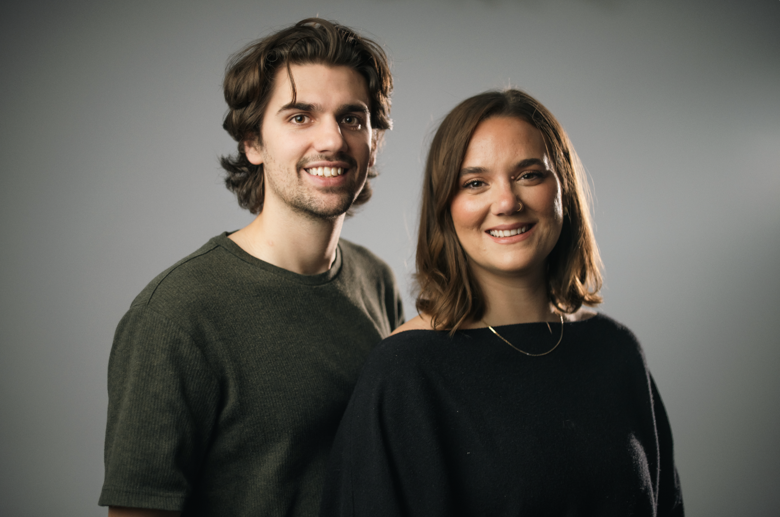 A young man with long, curly brown hair, and a young woman with long, wavy brown hair, smiling and posing together against a plain tan background.