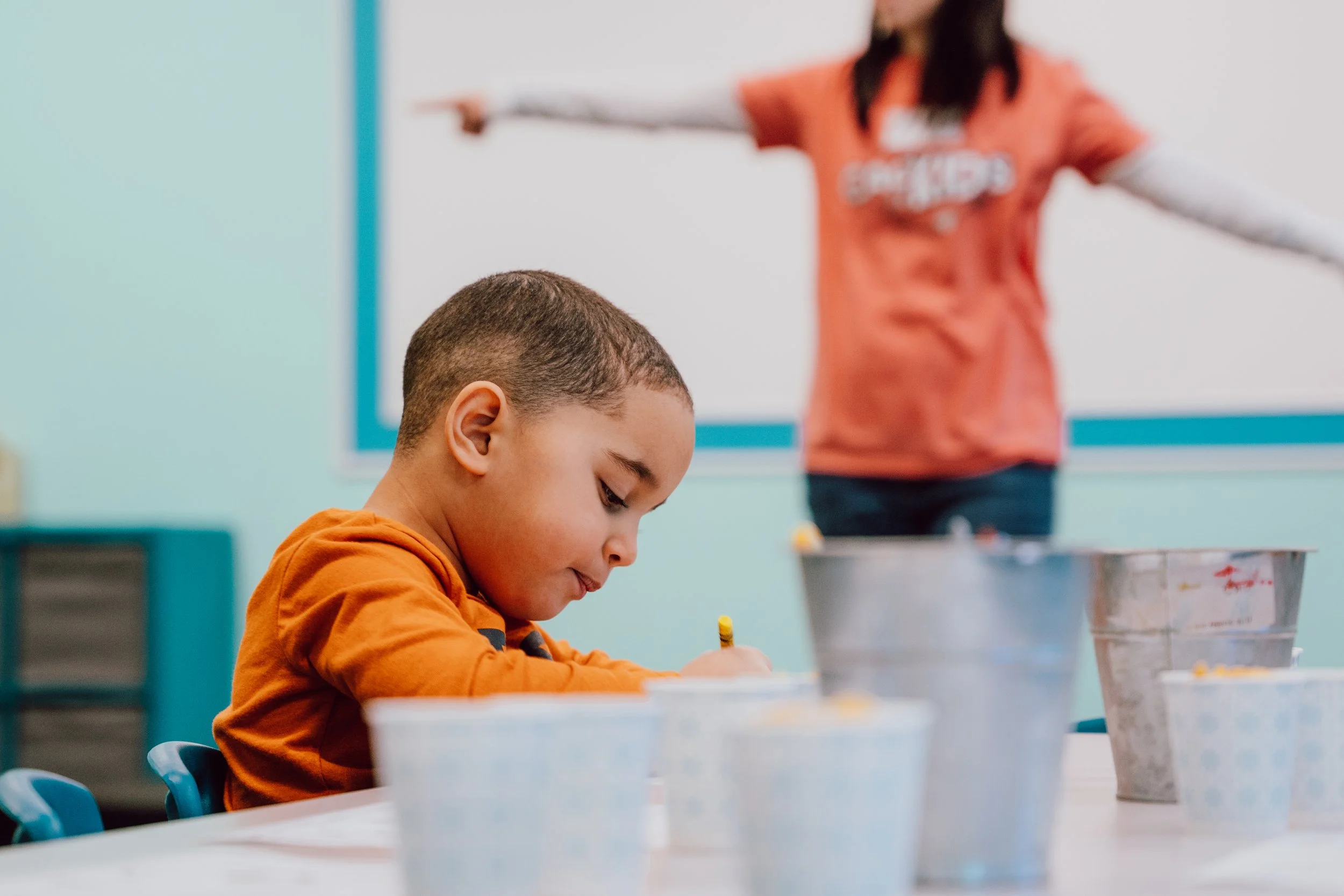 A young boy focused on writing at a table with buckets and containers nearby, while a woman in the background points and gestures towards the front of the classroom.