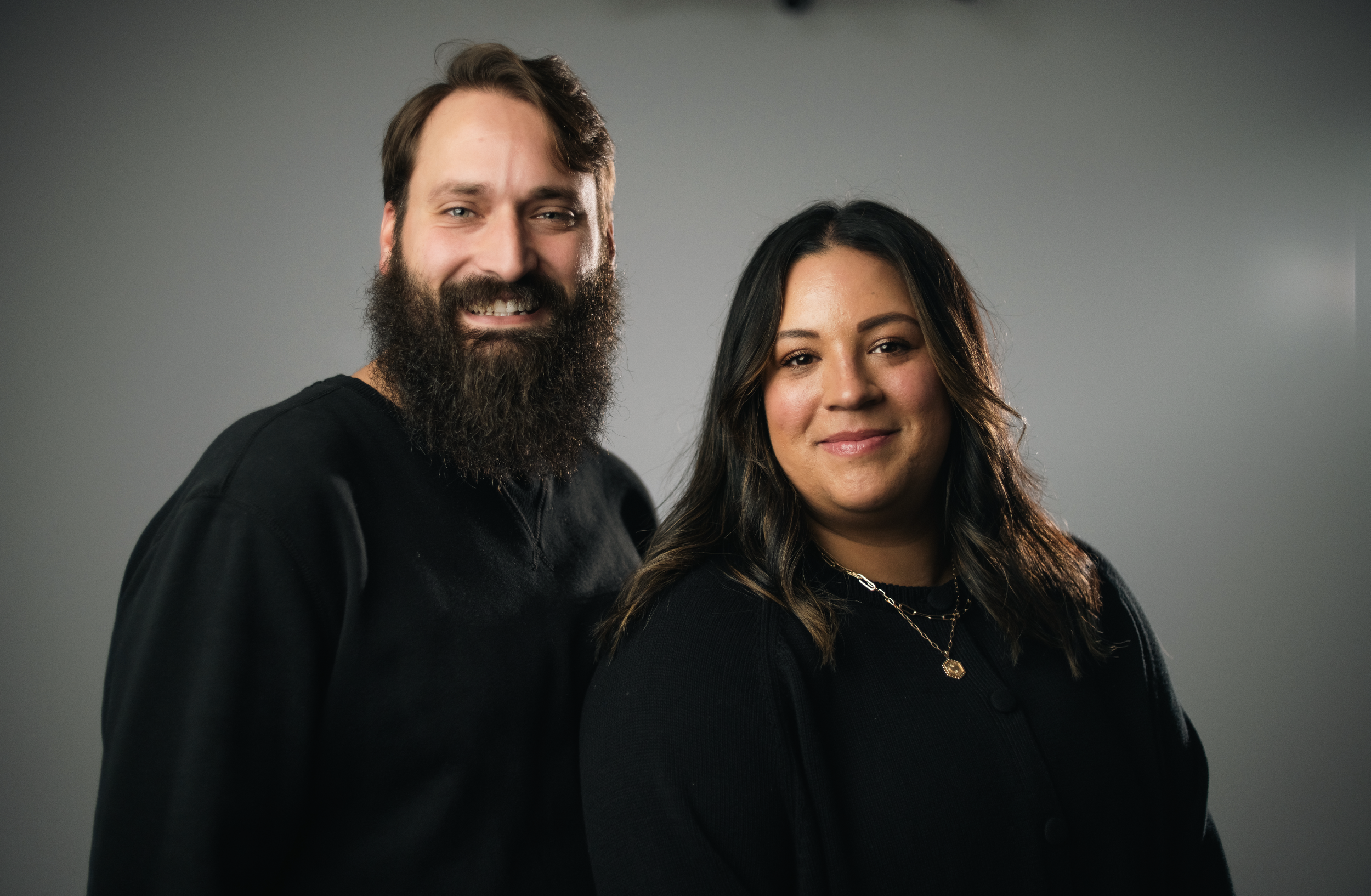 A smiling man with a beard and a smiling woman with dark hair posing together against a beige background.
