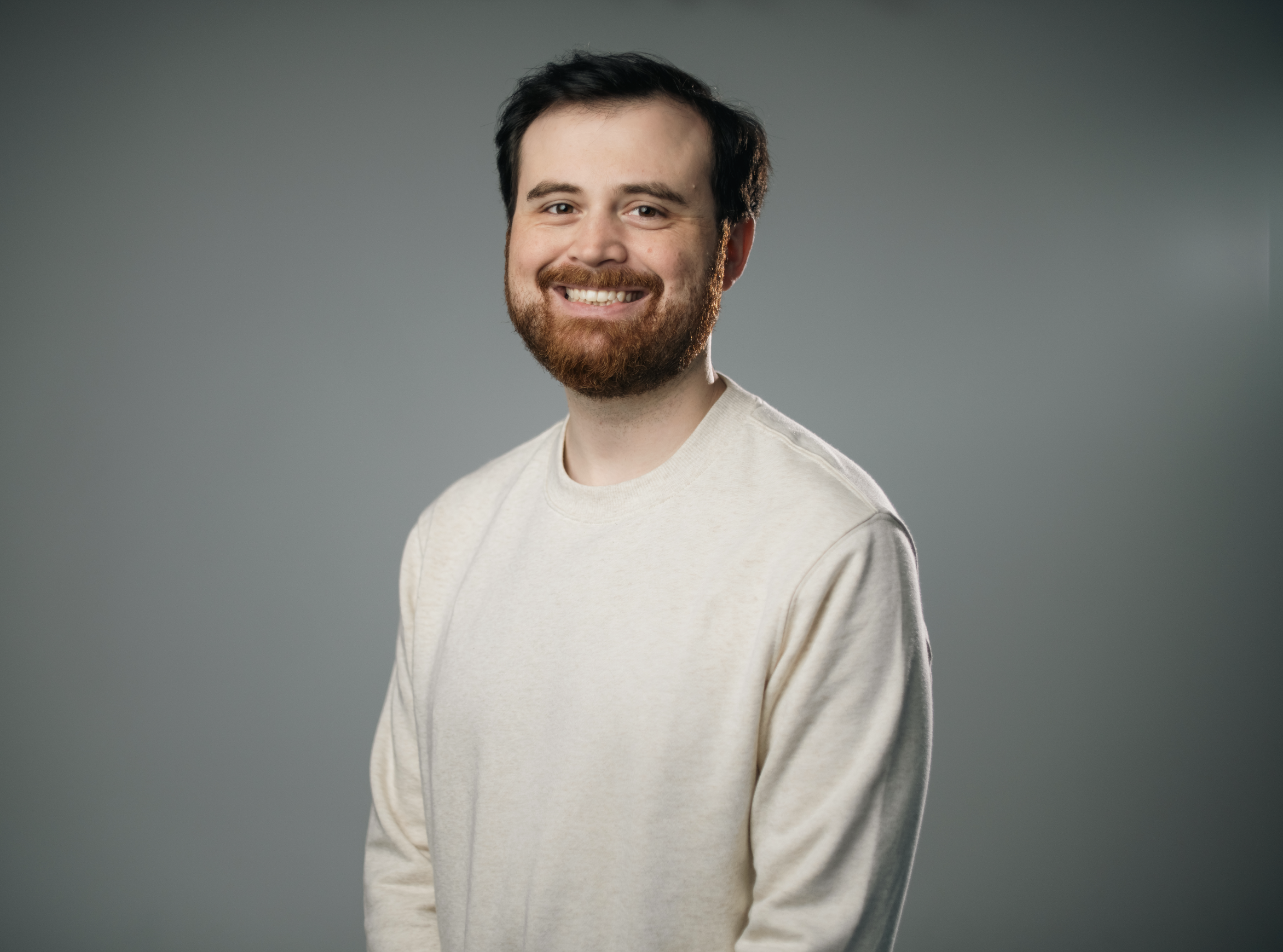 A smiling man with a beard and short dark hair, wearing a brown button-up shirt, standing against a plain beige background.