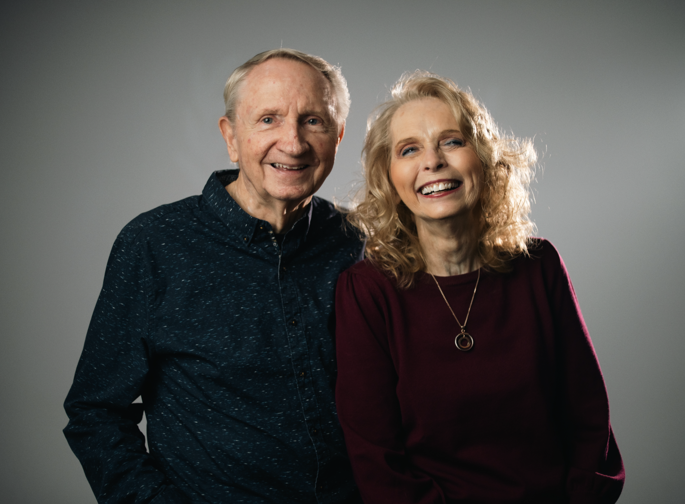 An elderly man and a woman stand together, smiling in front of a plain brown background.