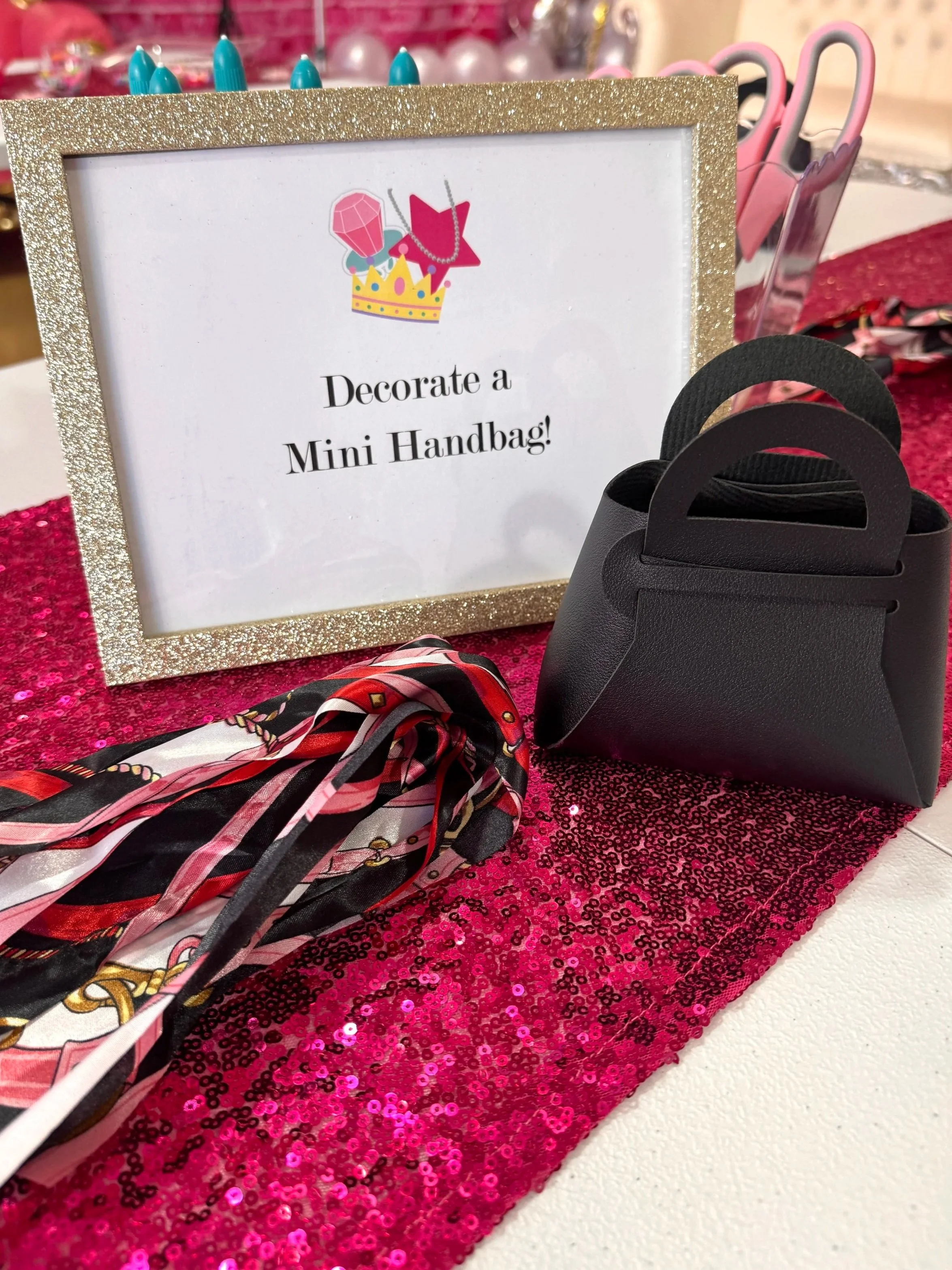 Sign with gold glitter border saying "Decorate a Mini Handbag!" surrounded by a black handbag, a colorful scarf, and pink sequined fabric.