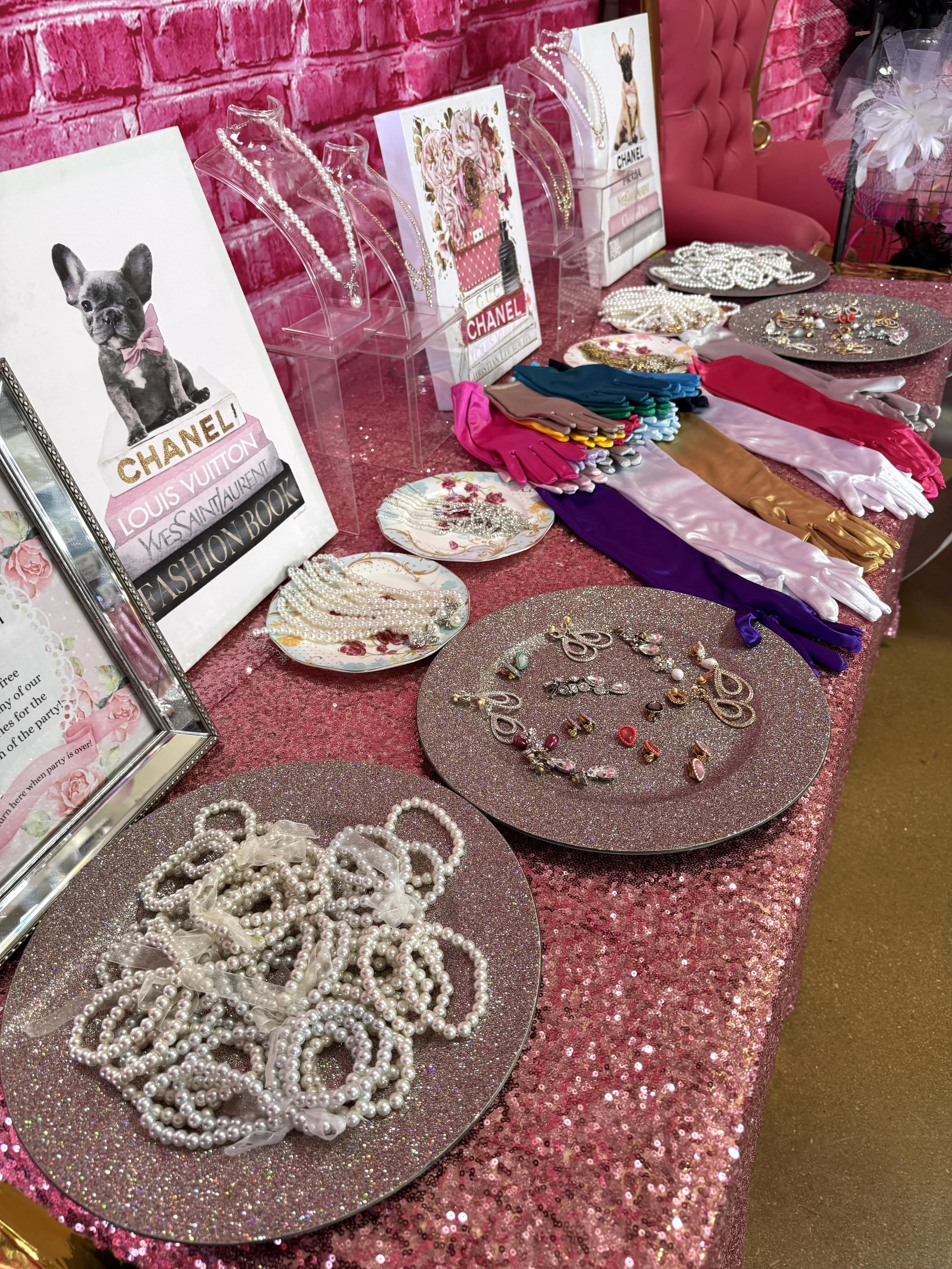 Display of jewelry including necklaces, bracelets, and earrings on sparkly plates and tables with a pink sequin tablecloth, in front of framed fashion book covers and artwork, against a pink brick wall.