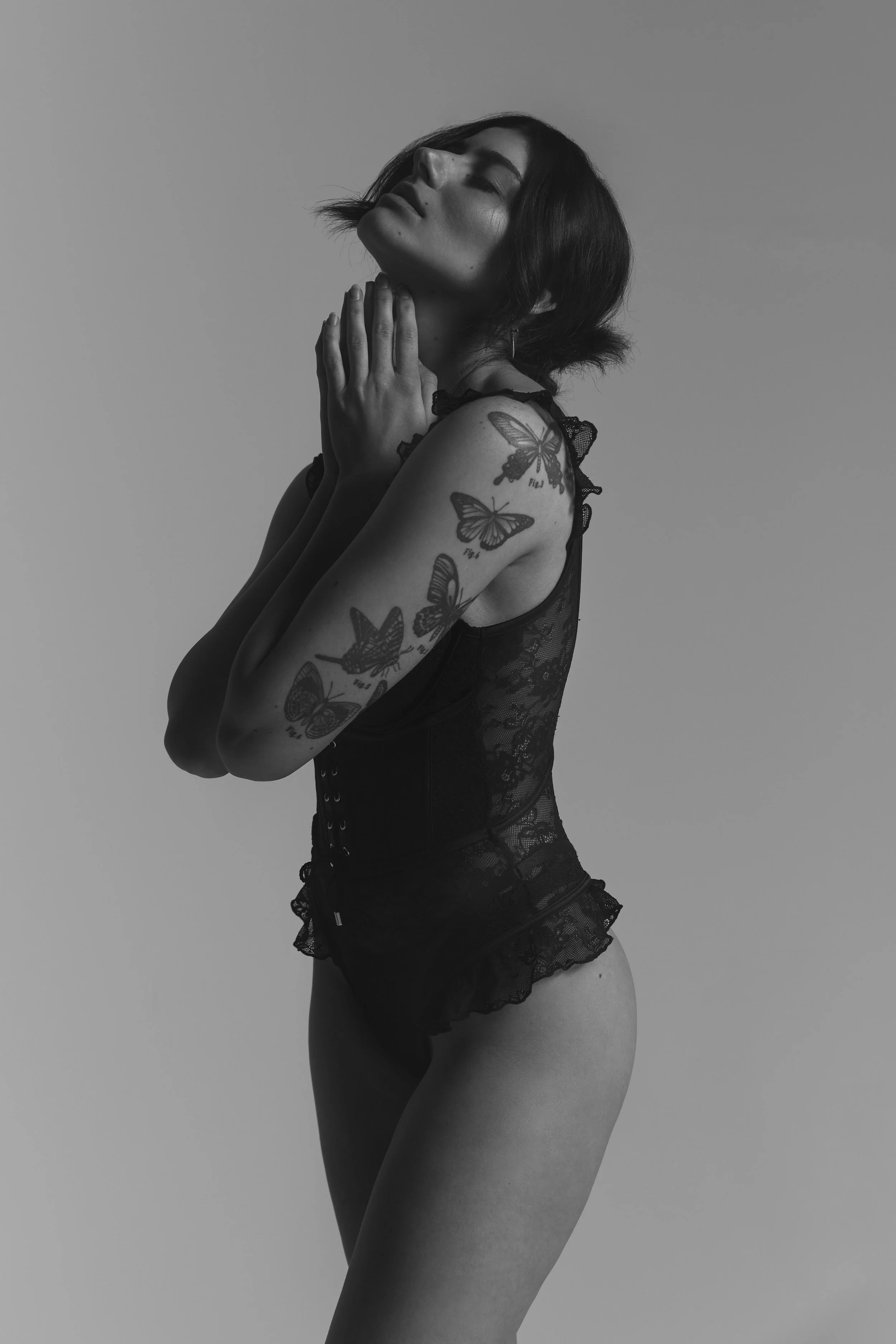 Black and white photo of a woman with short hair posing with her eyes closed, hands near her throat, wearing lace lingerie and displaying butterfly tattoos on her arm.