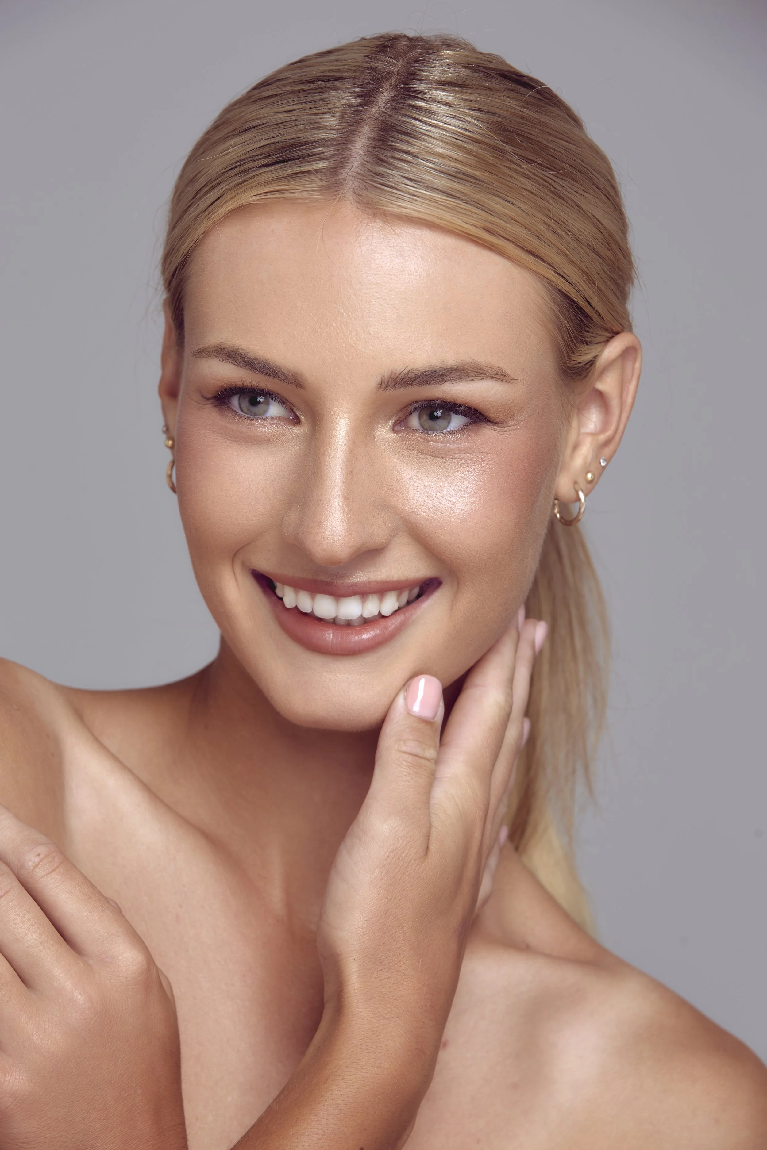 Close-up of a smiling woman with blonde hair, light skin, and multiple earrings, touching her face and shoulder, against a neutral background.