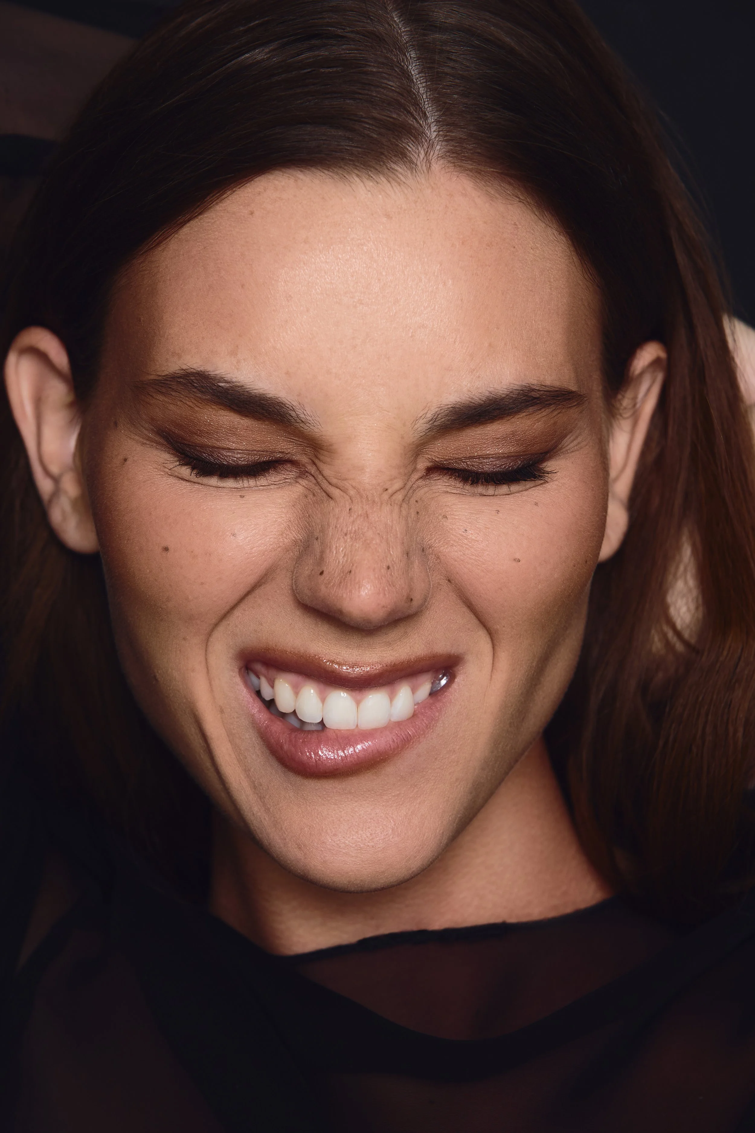 Close-up of a woman smiling with her eyes closed, showing her white teeth, brown hair, and wearing a black top.