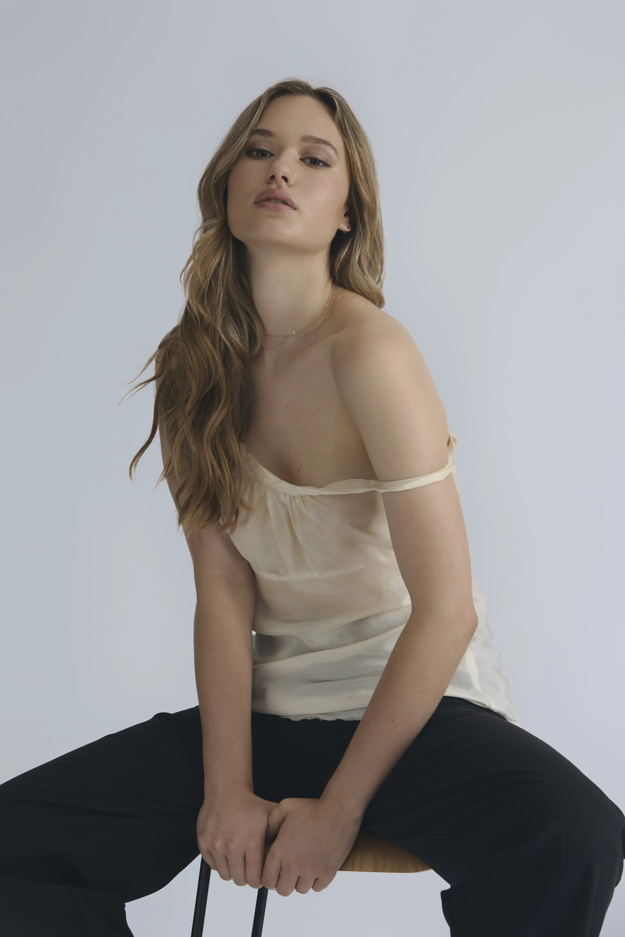 A young woman with long wavy brown hair, wearing a light-colored off-shoulder top and black pants, seated on a wooden chair against a plain light gray background.