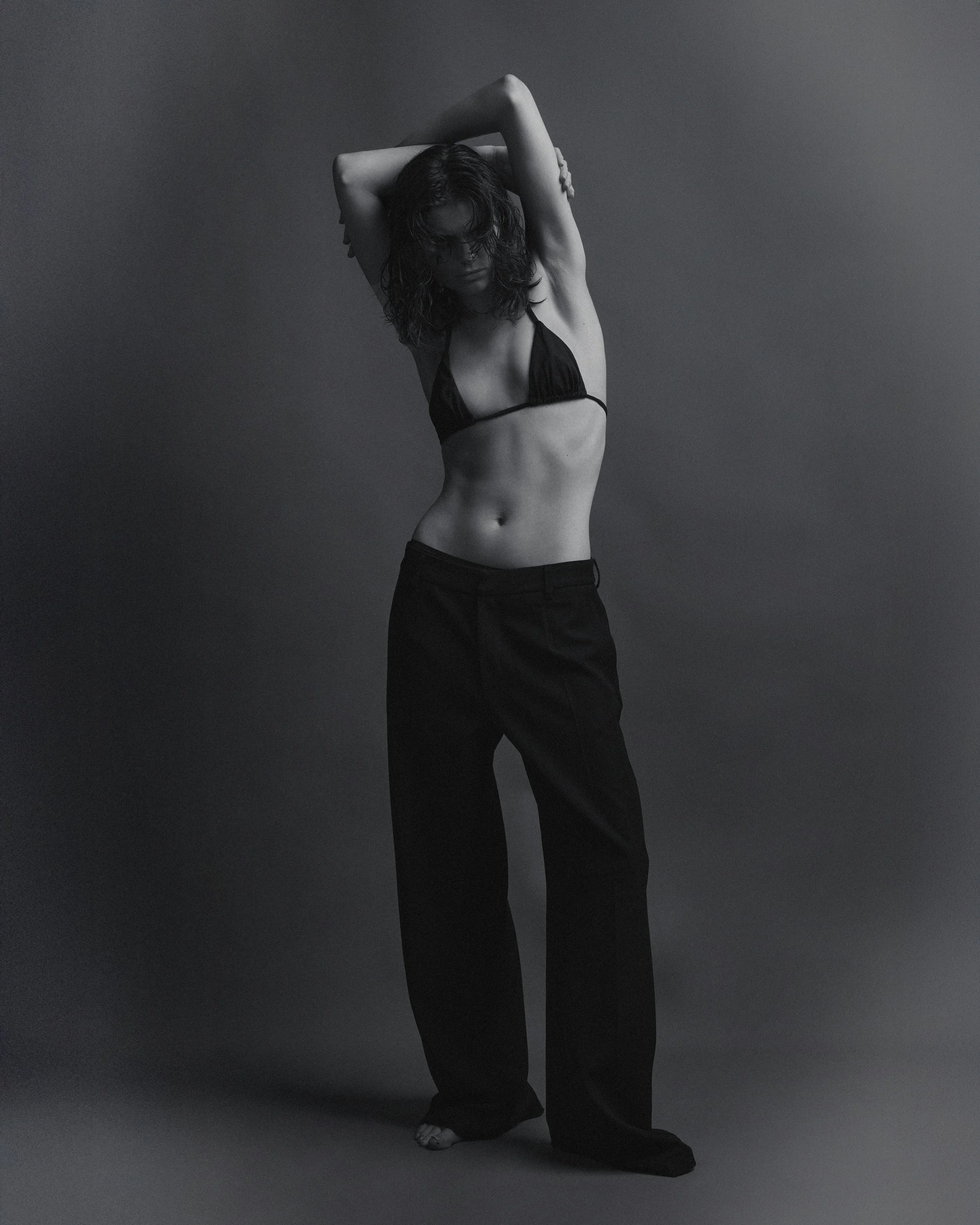 Black and white photograph of a woman with dark wavy hair posing with her arms raised, one hand resting on her head and the other along her back. She wears a black bikini top and loose black pants, against a plain, dark background.