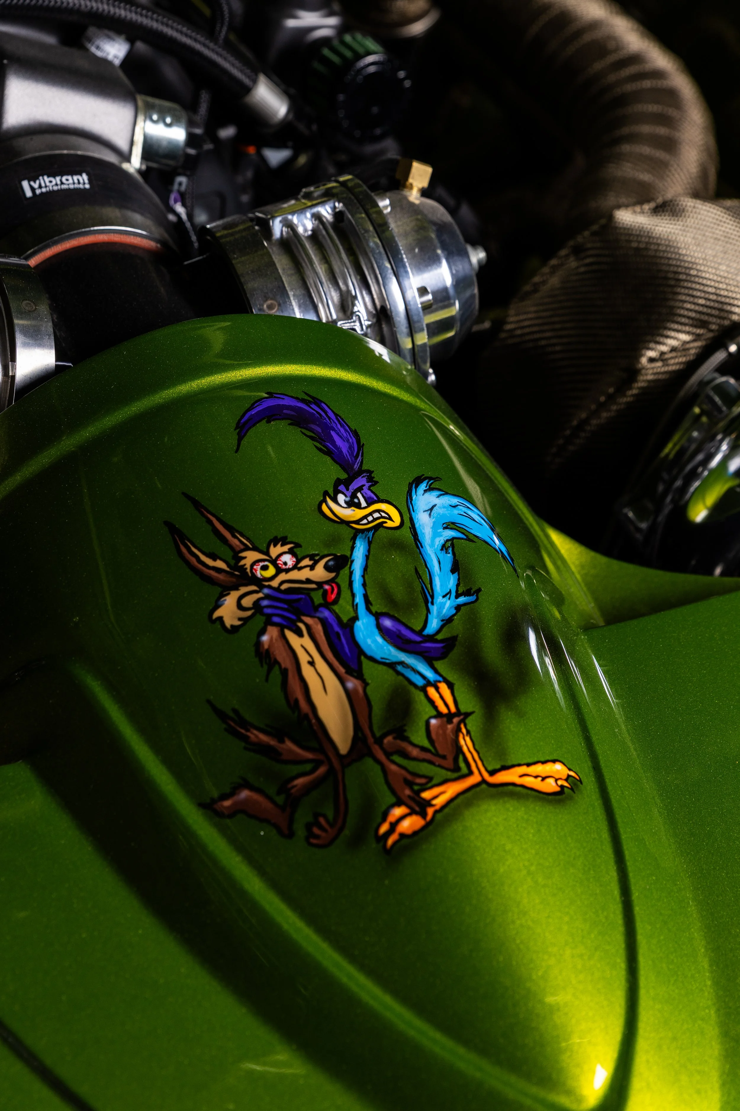 Close-up of a green motorcycle fuel tank featuring a cartoon decal of Wile E. Coyote and the Road Runner.