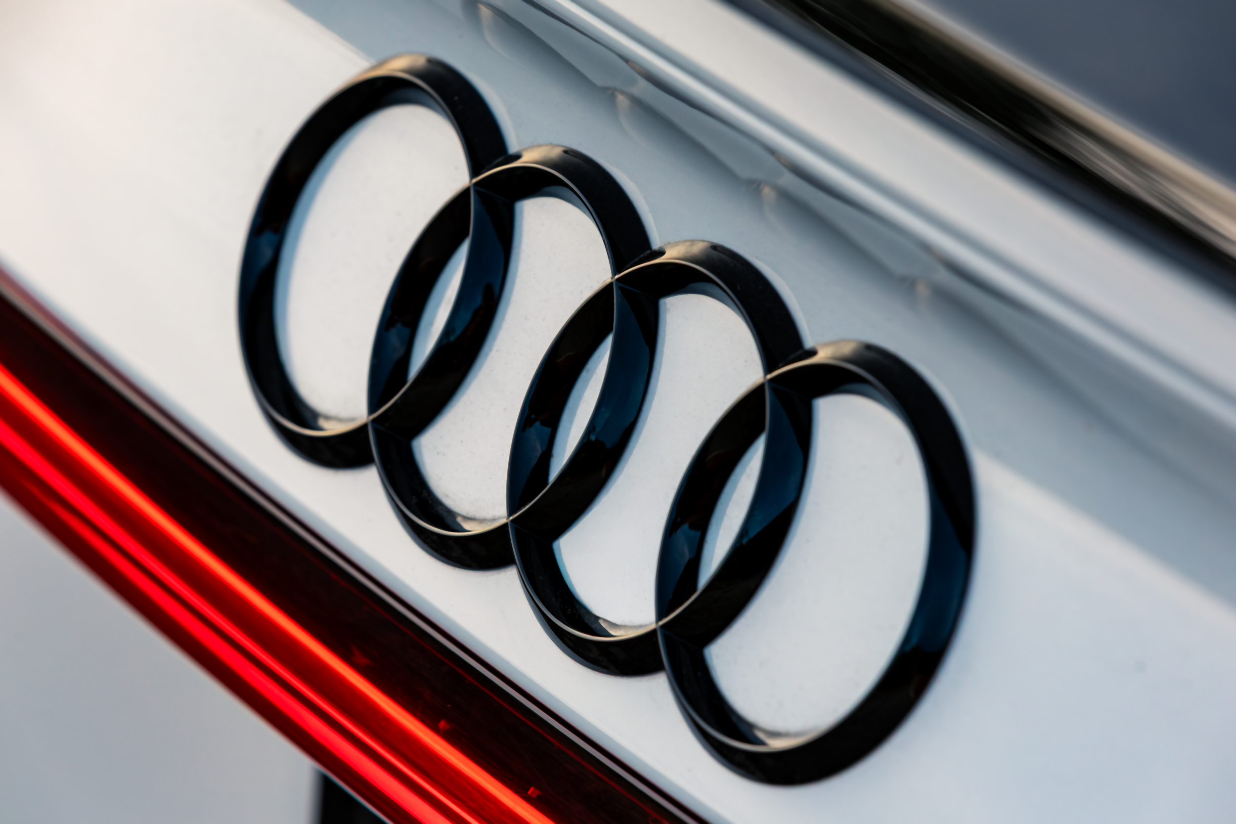 Close-up of four black Audi logo rings on the back of a silver car, with a red taillight visible in the bottom left corner.