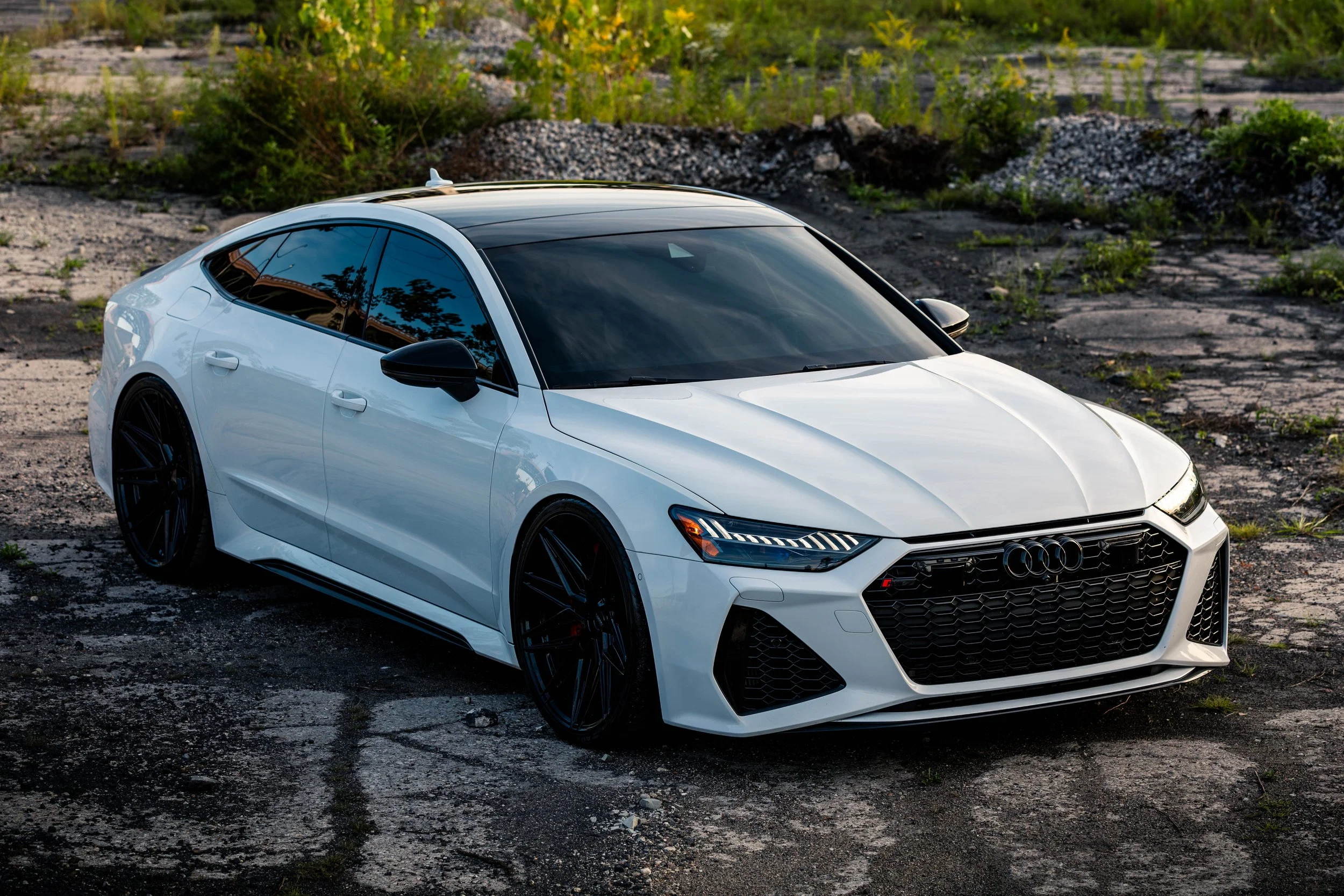 White Audi sporty hatchback car parked on a cracked asphalt surface with green bushes in the background.