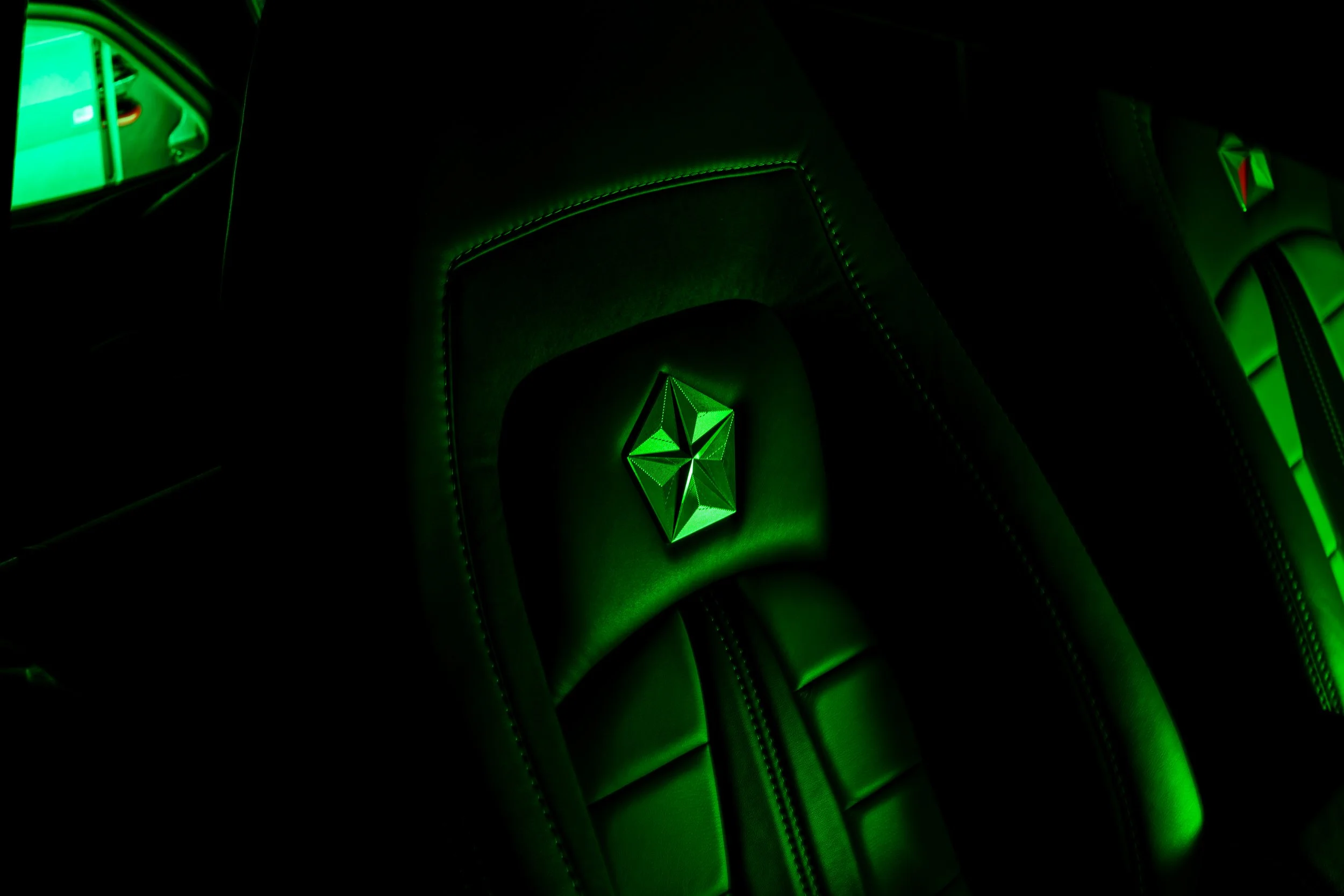 Close-up of a black car seat with illuminated green logo, with reflections of green lighting on the leather surface.