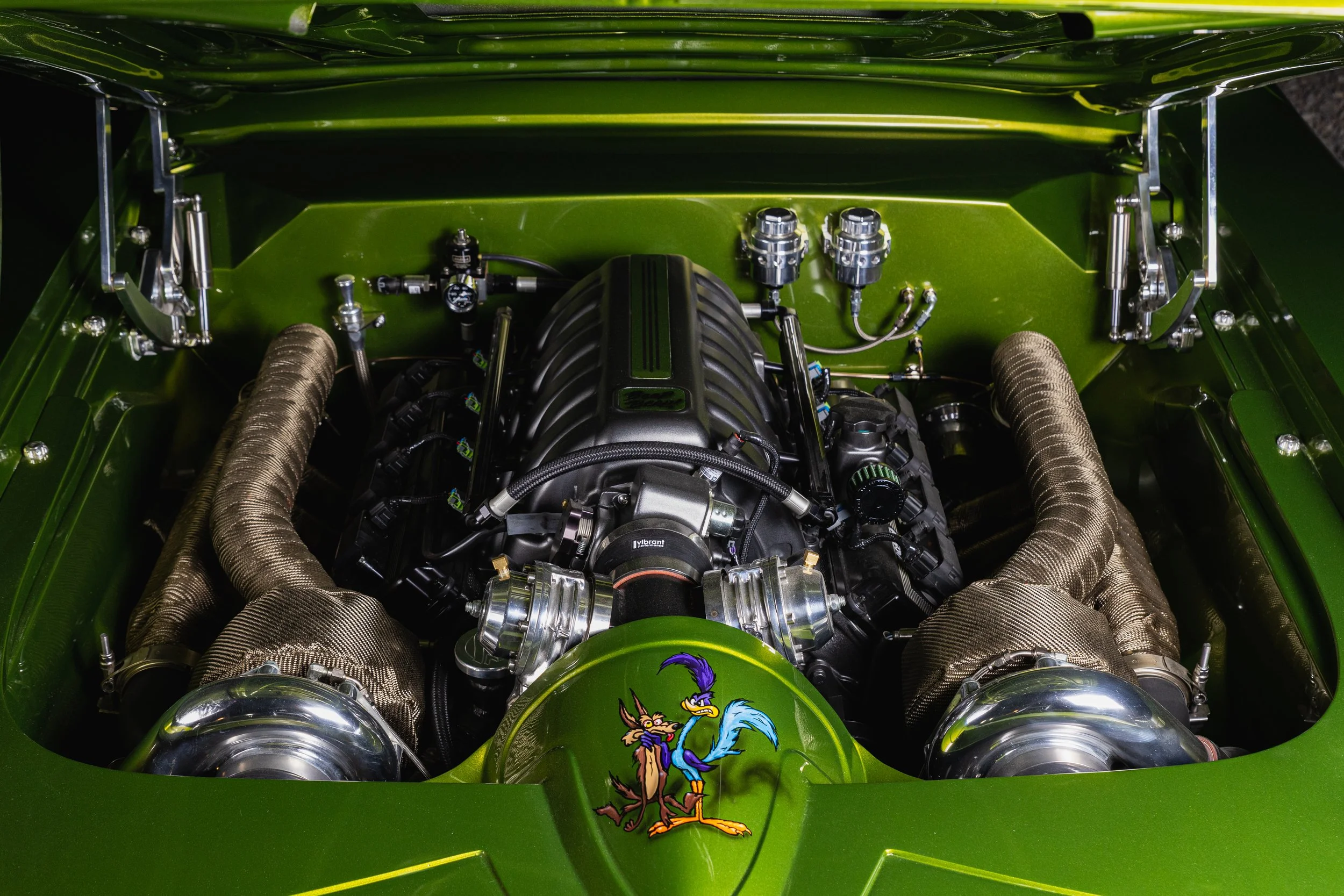 Close-up view of a bright green car engine with a cartoon rooster and dog decal on the hood, showing intricate mechanical parts and metallic exhaust pipes.
