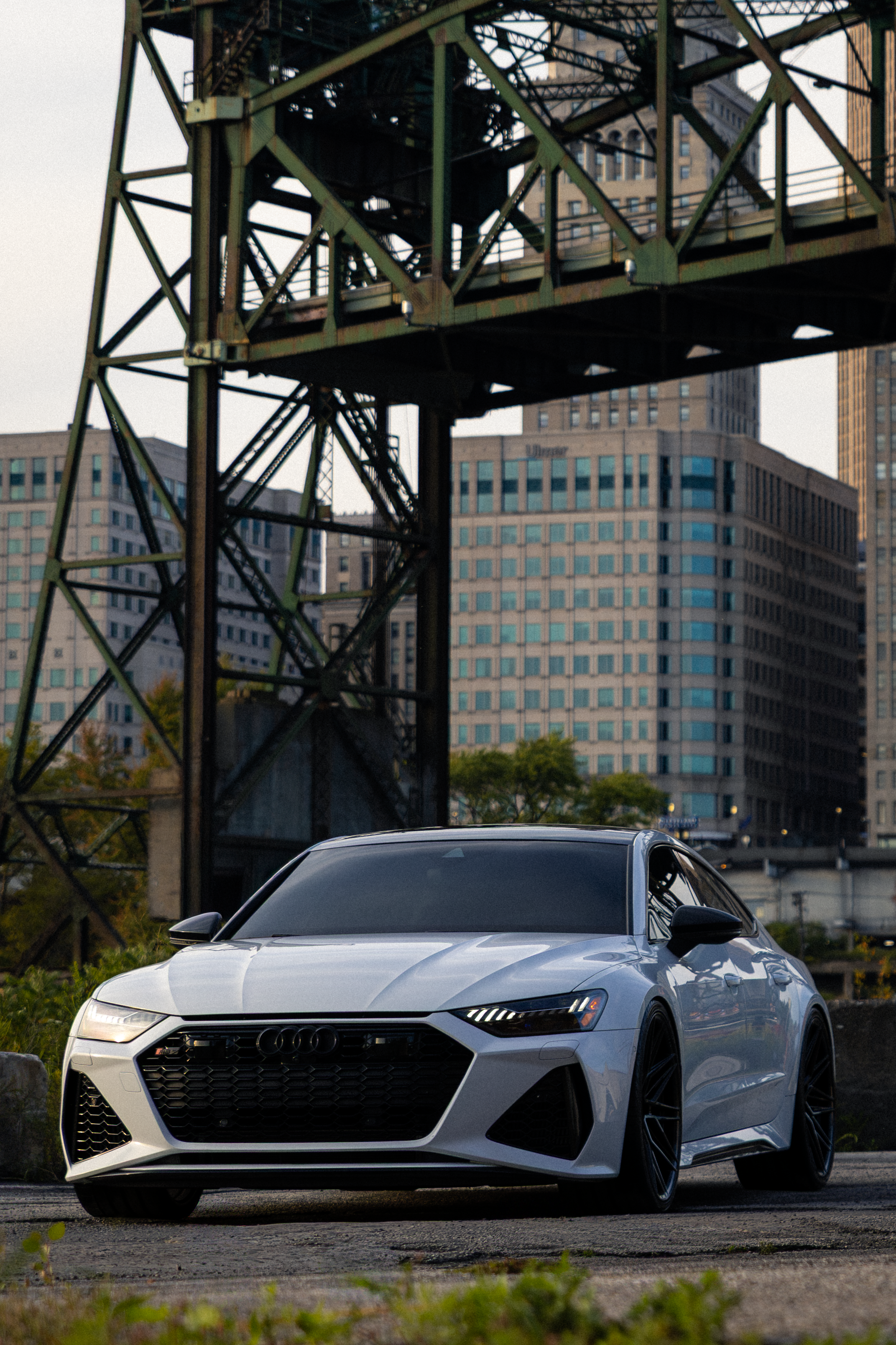 A white Audi sports car parked outdoors with a metal bridge structure and city buildings in the background.