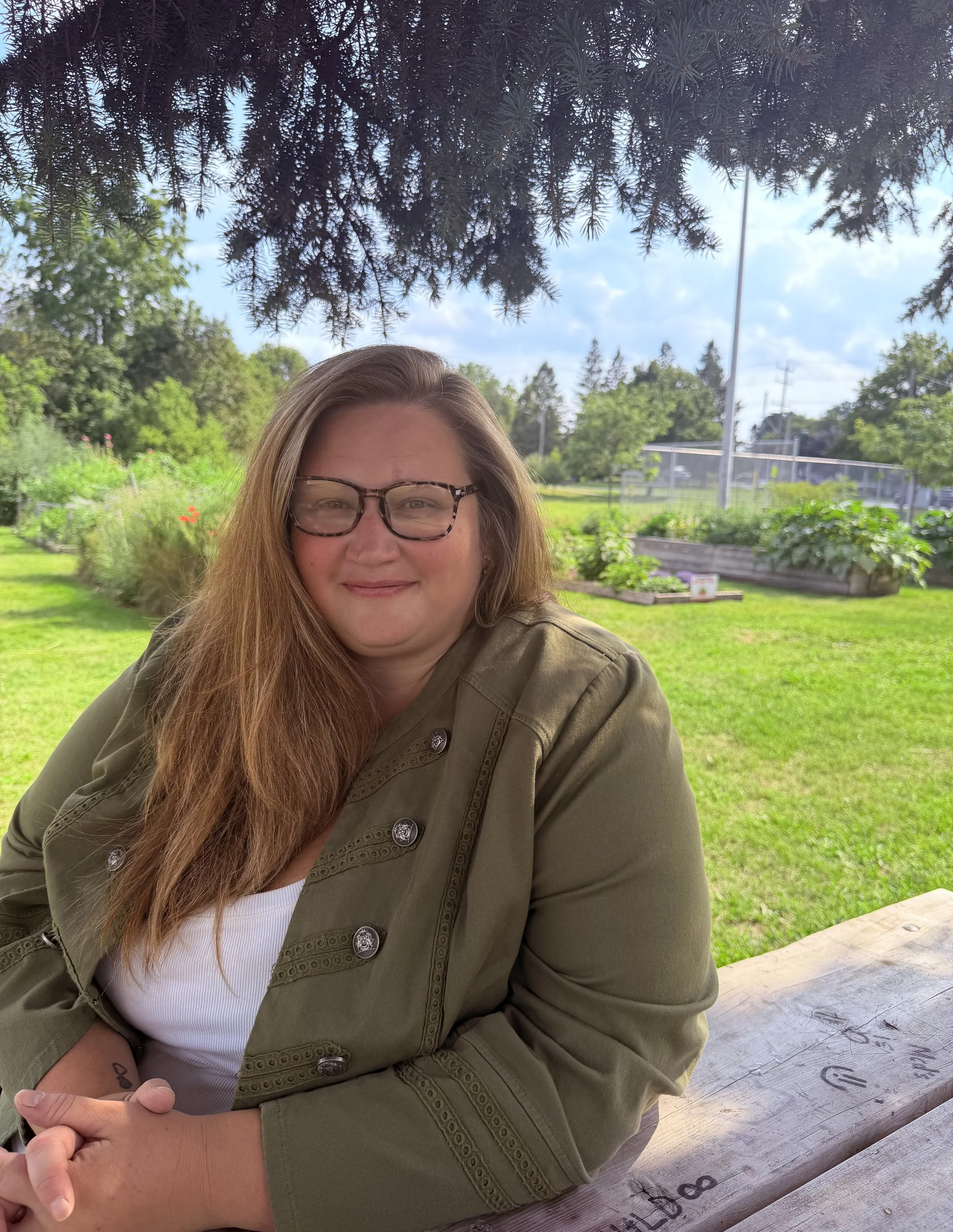 A woman with long brown hair, glasses, and a green jacket smiling outdoors in front of trees and a blue sky, with garden beds and a tennis court in the background.