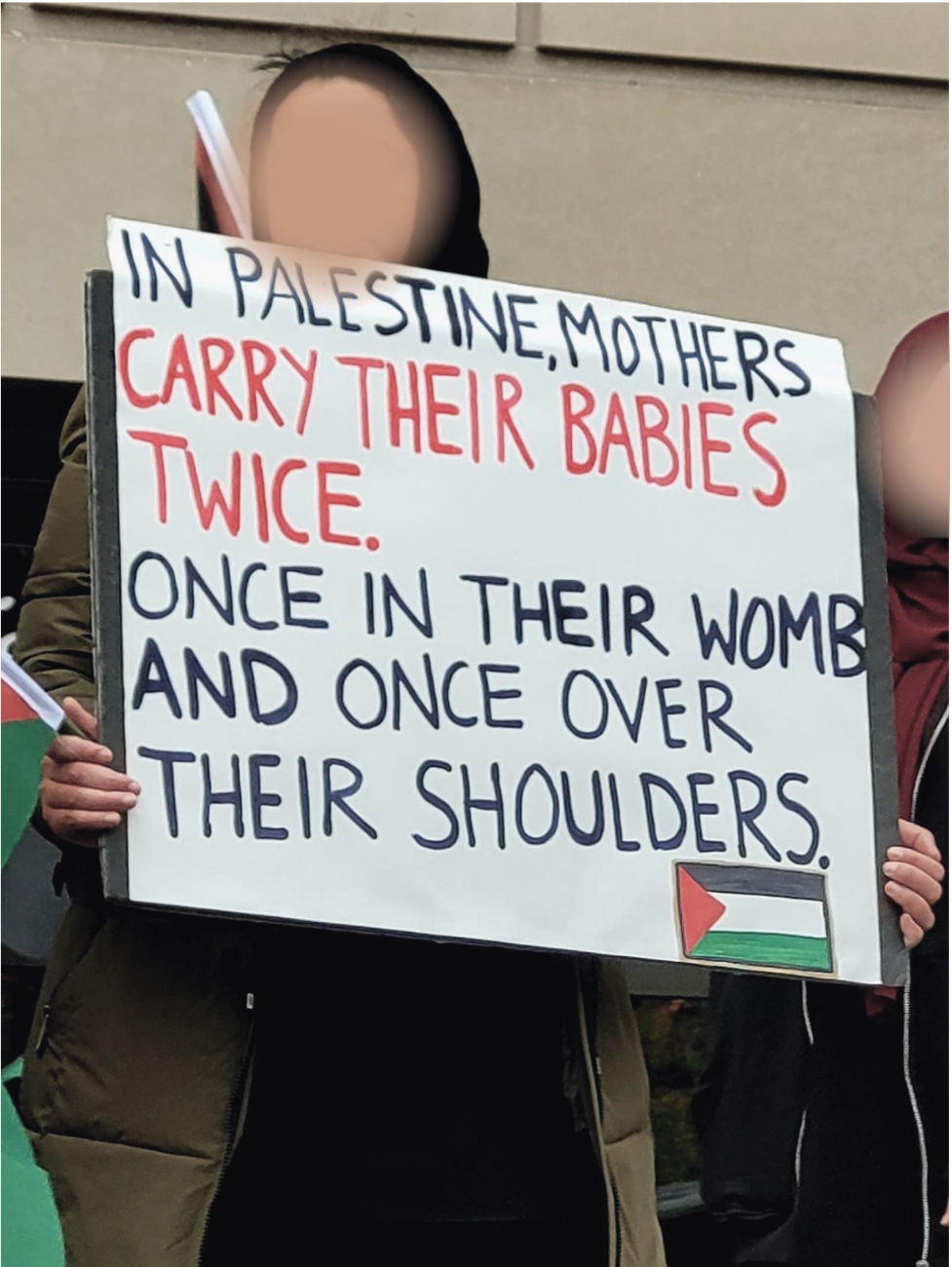 Person in a long winter coat and hijab holding a large white sign with black and red lettering and a Palestinian flag that says, "In Palestine, mothers carry their babies twice. Once in their womb and once over their shoulders."