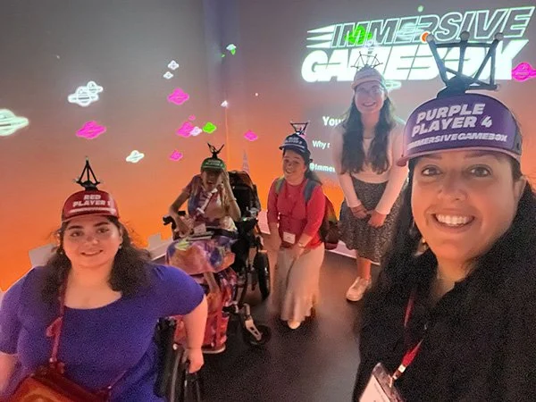 Four women, two in wheelchairs, at an immersive gaming event with a large screen behind them displaying the words 'IMMERSIVE GAME' and colorful graphics.