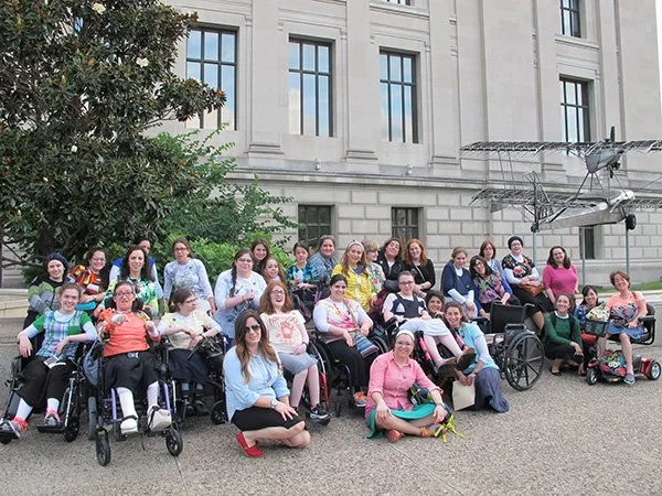 Group of women, some in wheelchairs, posing outdoors in front of a historic building with a large sculpture of an airplane hanging above them.