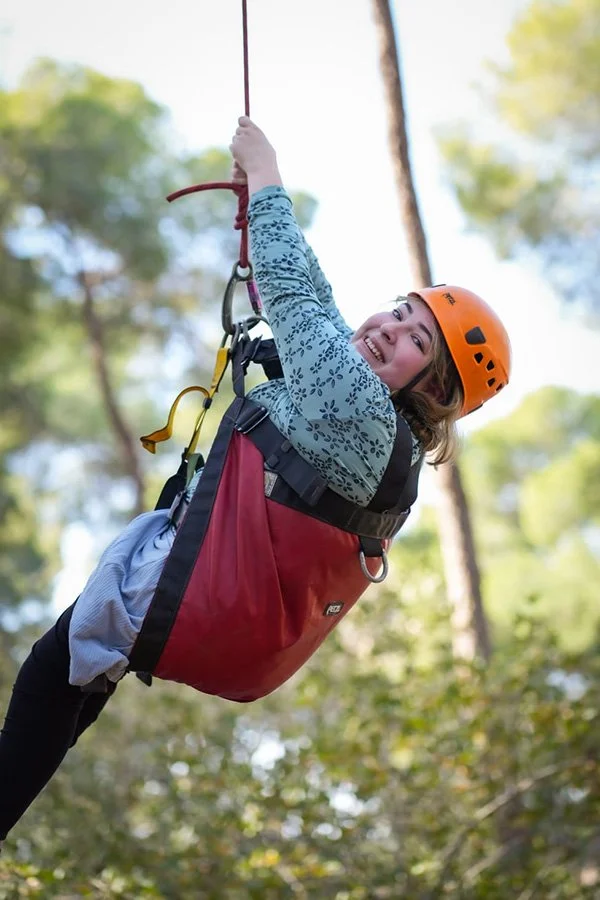 A woman with a helmet smiling while zip-lining through a forested area.