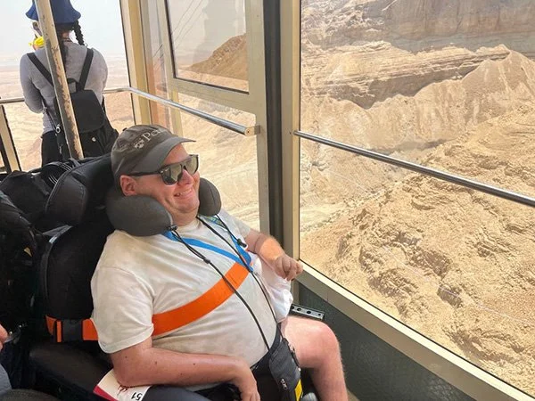 A person with a physical disability sitting in a wheelchair inside a glass-enclosed observation area at the Grand Canyon, looking out at the canyon landscape.
