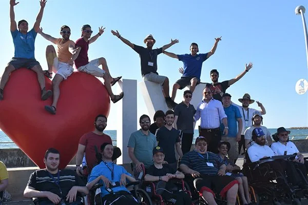 Group of people with disabilities and supporters, some in wheelchairs, gathered outdoors around large red heart-shaped sculpture with the letters 'LOVE' in the background, smiling and raising their hands.