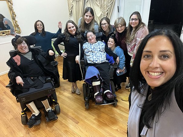 Group of nine women and two individuals with disabilities, one in a wheelchair, smiling and posing for a selfie in a room with wooden floors and elegant curtains.