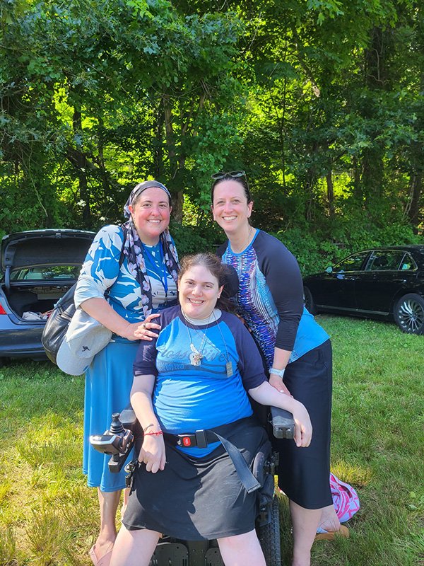 Three women outdoors in a grassy area with trees in background, two standing and one seated in a motorized wheelchair, all smiling.