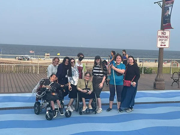 Group of people, including children and adults, posing on a striped blue awning near the beach, with the ocean and a no parking sign in the background.