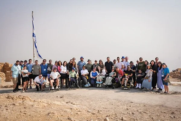 Group of people, some using wheelchairs, gathered outdoors with a Greek flag on a pole and rocky terrain in the background.
