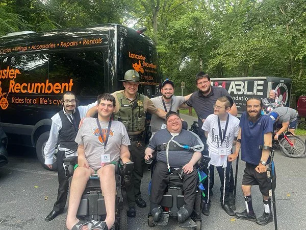 Group of eight people, including two in wheelchairs, outdoors near black and pink vehicles with trees in the background.