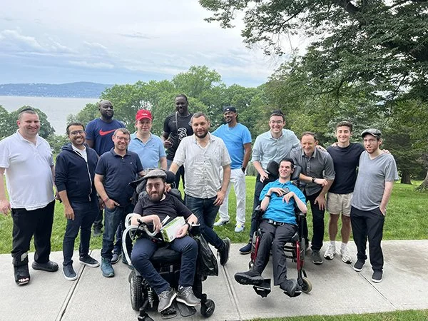 Group of fifteen men outdoors on a grassy area with trees and a body of water in the background. Some are standing, two are sitting in wheelchairs in the front. All are smiling and dressed casually.