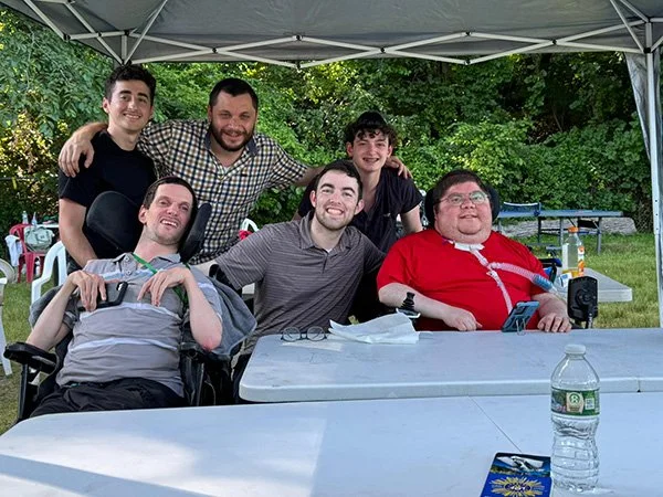 Group of six people, including one in a red shirt with a wheelchair, smiling and gathered under a canopy at an outdoor gathering with tables and greenery in the background.