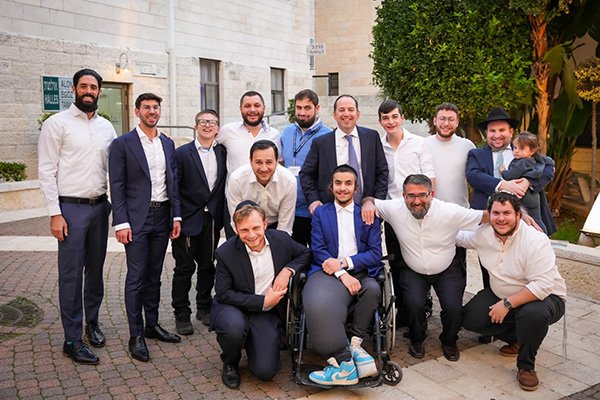 Group of fifteen men, some in suits, posing outdoors in front of a light-colored building with greenery.