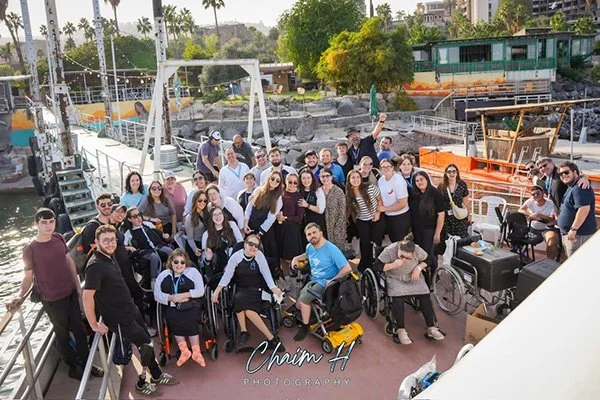 Group of people on a boat, some in wheelchairs, gathered together for a photo with a city and trees in the background.