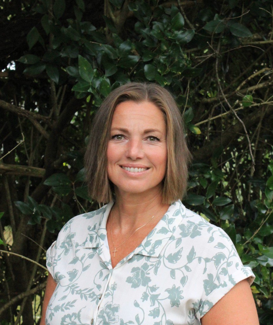A woman with shoulder-length brown hair, smiling, standing outdoors in front of green leafy bushes, wearing a white blouse with a light gray floral pattern and a gold cross necklace.