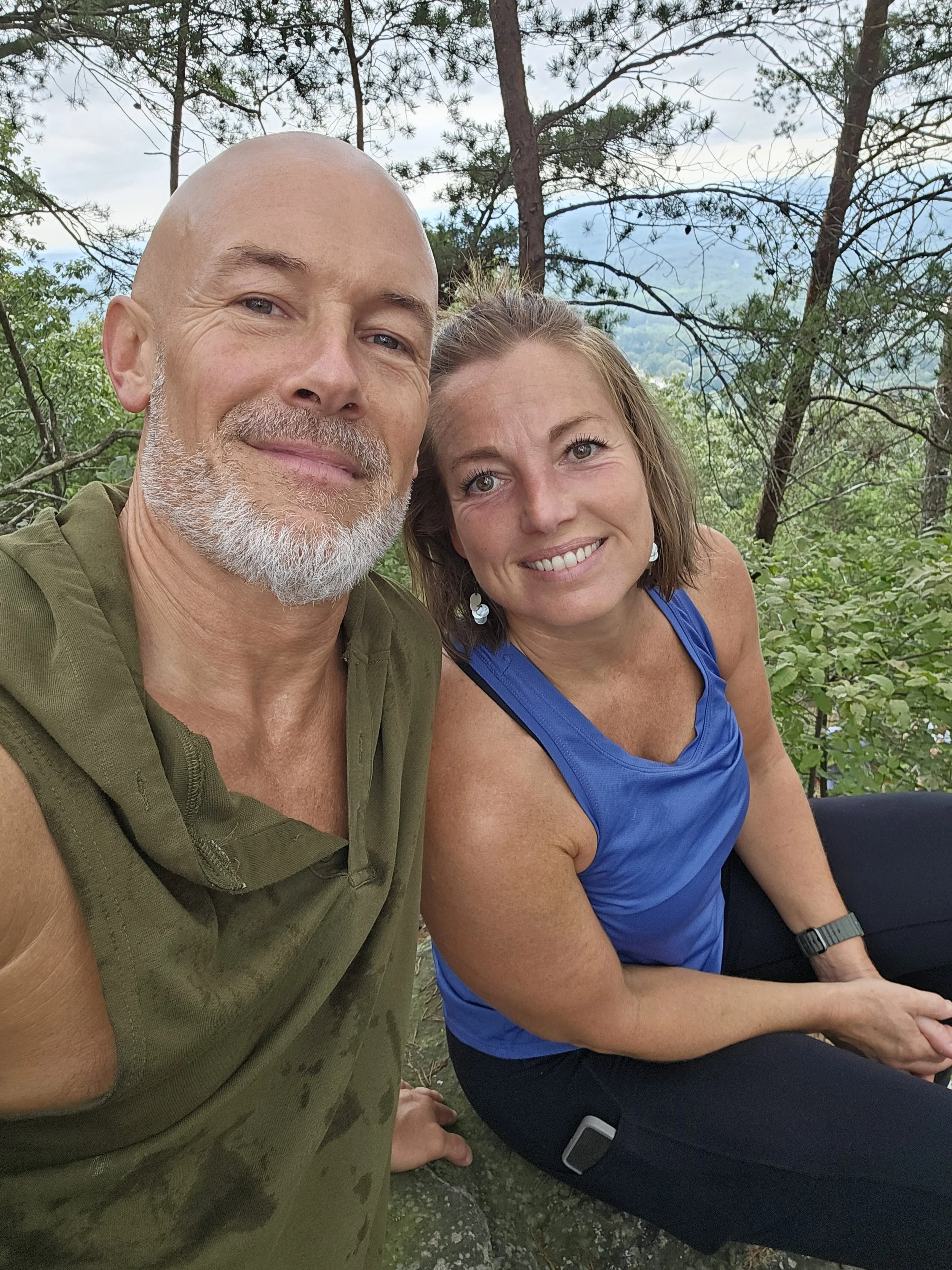 A man and woman taking a selfie outdoors in a forested area with trees and mountains in the background. They are smiling, with the man wearing a sleeveless green shirt and the woman in a blue tank top.