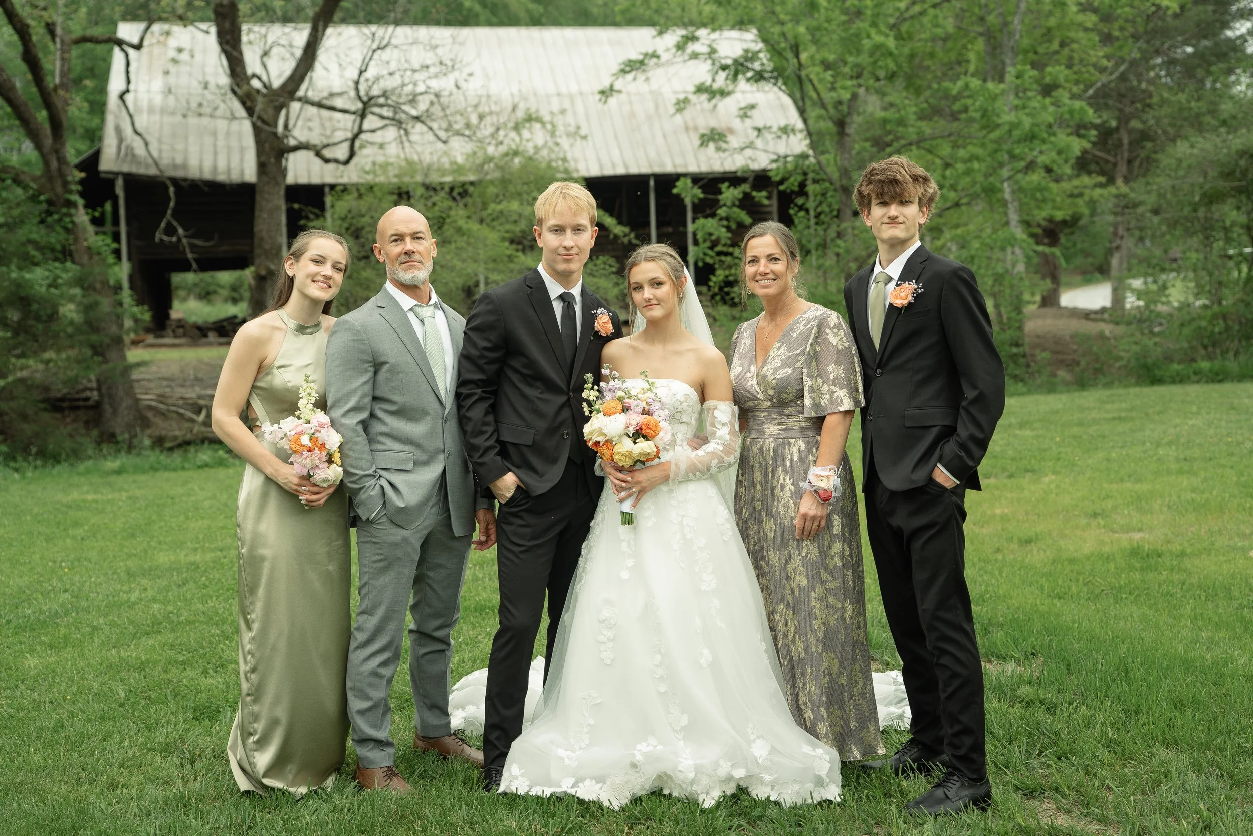 A wedding photo featuring a bride in a white gown holding a bouquet, standing with five people outdoors on a grassy area with trees and a large barn in the background. The group includes a groom, two women, and two men, all dressed in formal attire.
