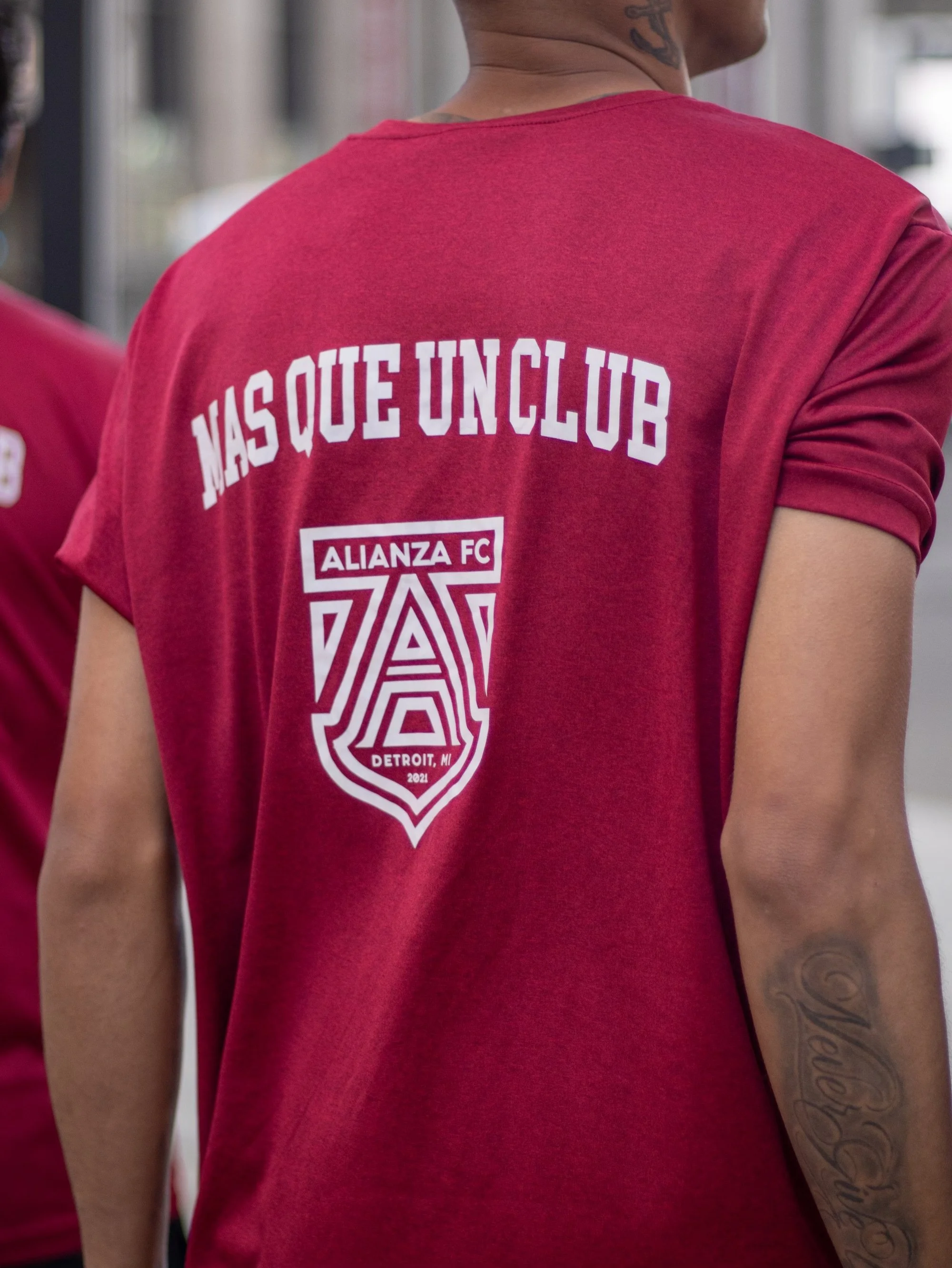 Back view of a person wearing a red T-shirt with white text and logo saying "MAS QUE UN CLUB, ALIANZA FC, DETROIT, MI".