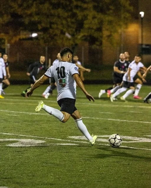 A soccer player wearing a white jersey with number 10 on the back, kicking a soccer ball on a field during a match at night. Other players are visible in the background. At an Alianza FC game in Detroit, MI.