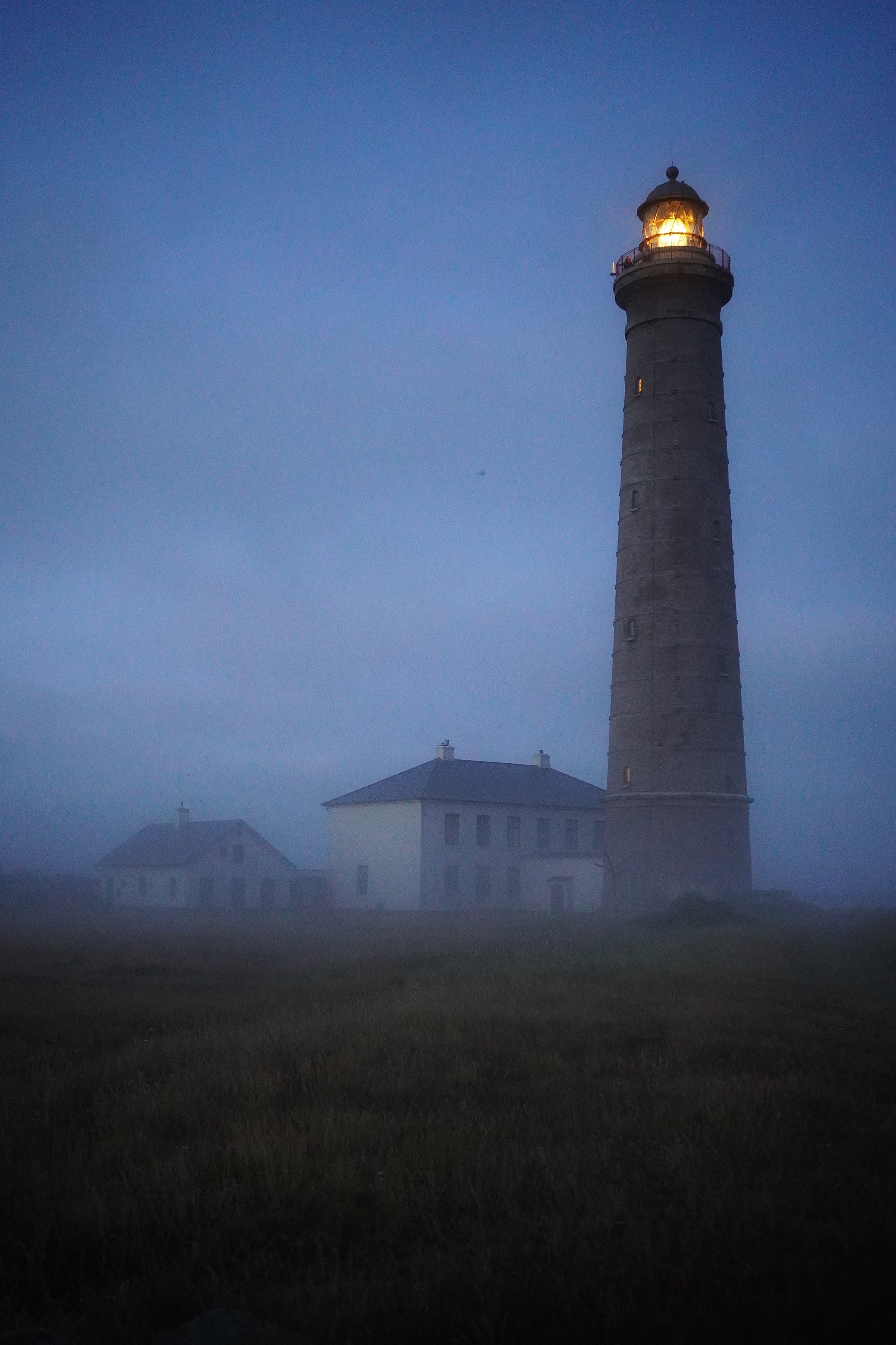 Lysende fyrtårn ved skumring eller tåge, omgivet af to hvide huse, i en landlig område.