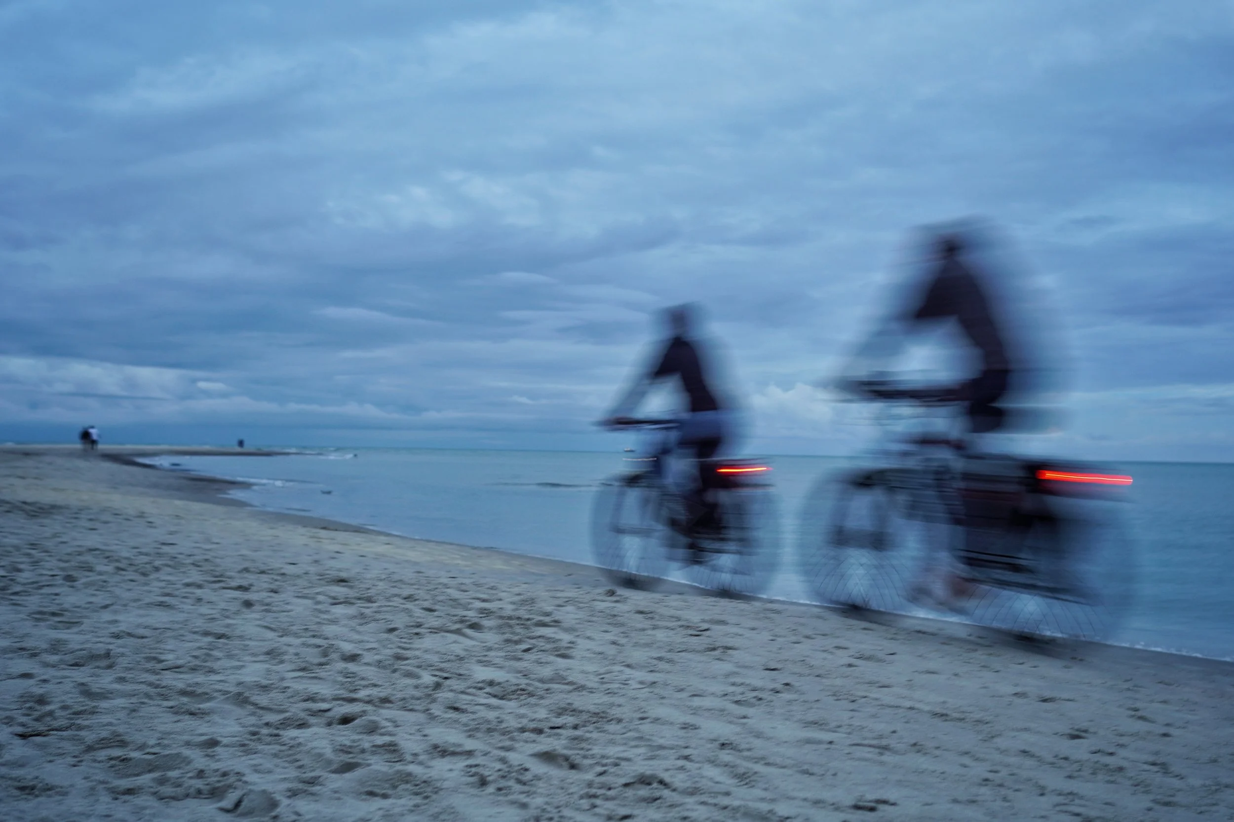 To personer på cykler ved stranden i skumringstiden, med blødt skydække og stillestående vand.