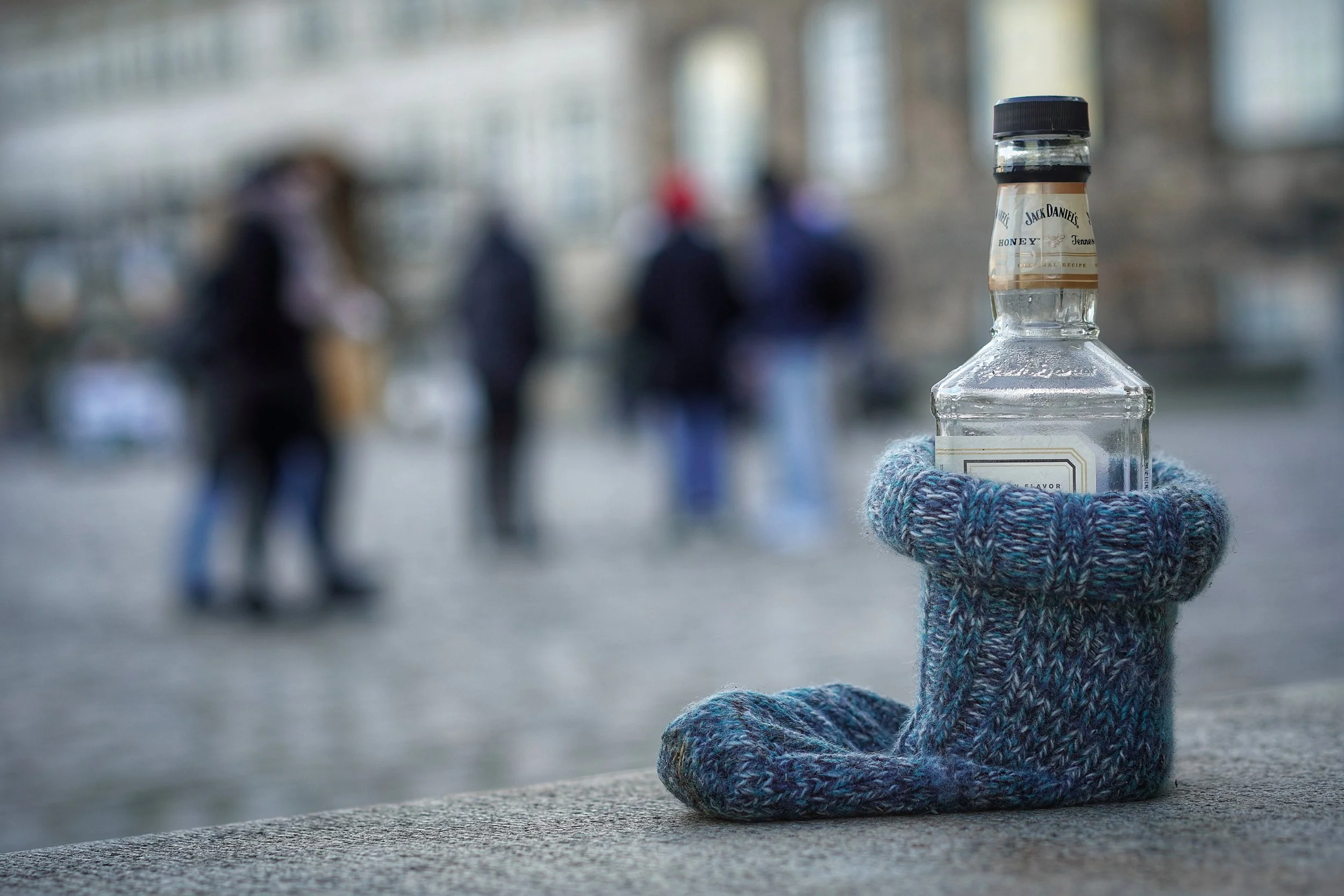 En klar glasvodkaflaske med en håndenstrikket sok, der holder det stående. I baggrunden er der tre personer, der går på en bro eller gangbro.
