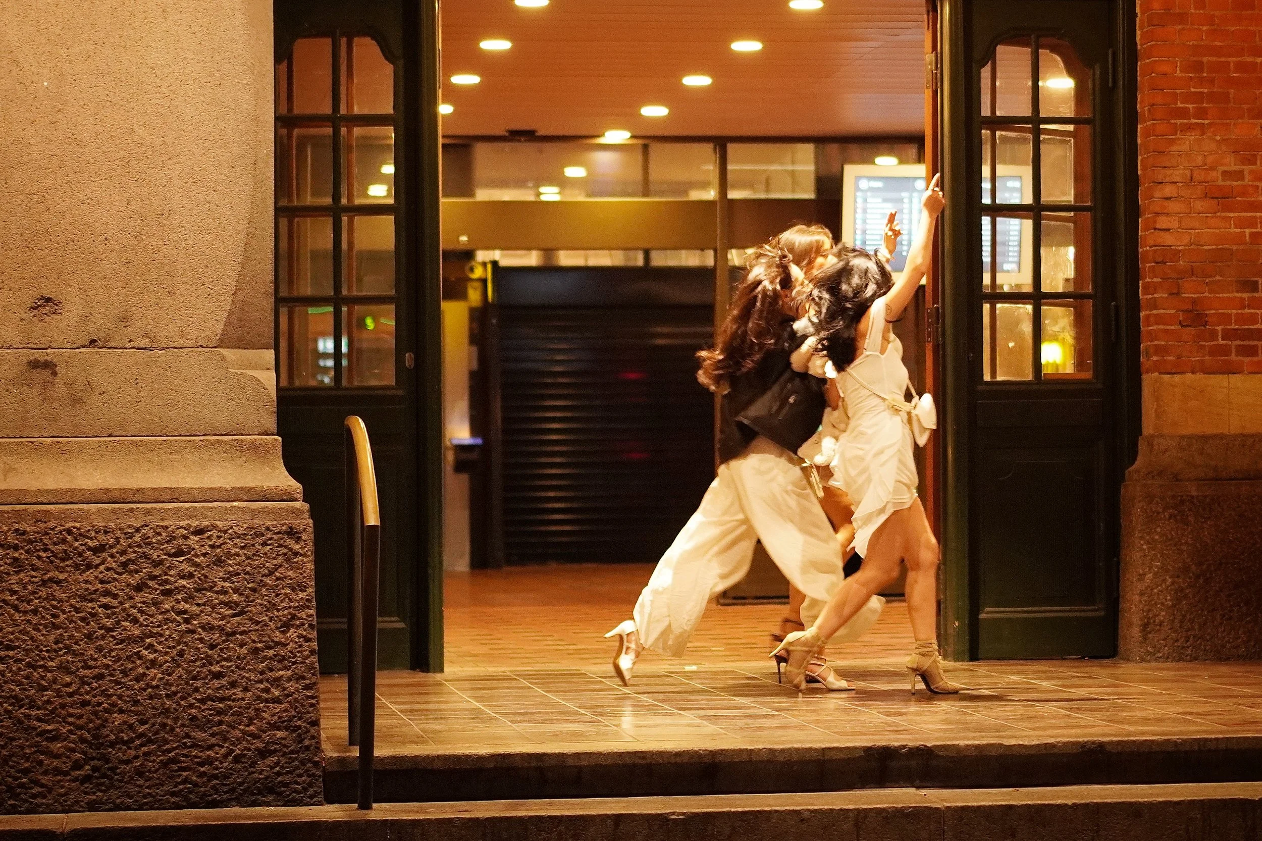 Tre kvinder går ind i en bygning gennem en åben dør, iført kjoler og skyhøje hæle, i en urban, velbelyst setting om aftenen.