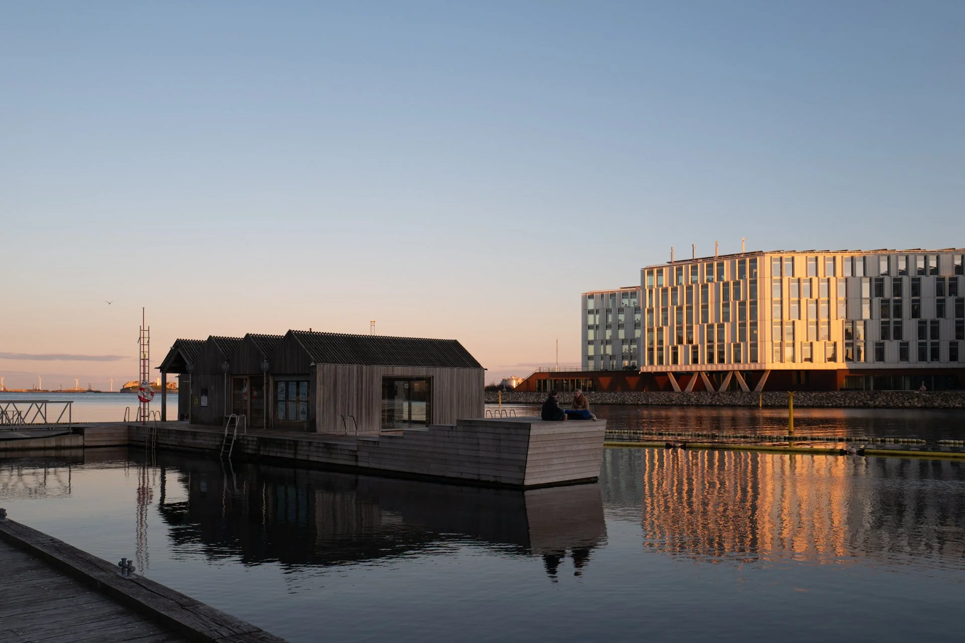 Moderne havnebad og kontorbygninger i solnedgang ved havnefronten. Aftenlys reflekterer i vandet ved et moderne havnebad – et billede af arkitektur, ro og byliv ved vandet i København.