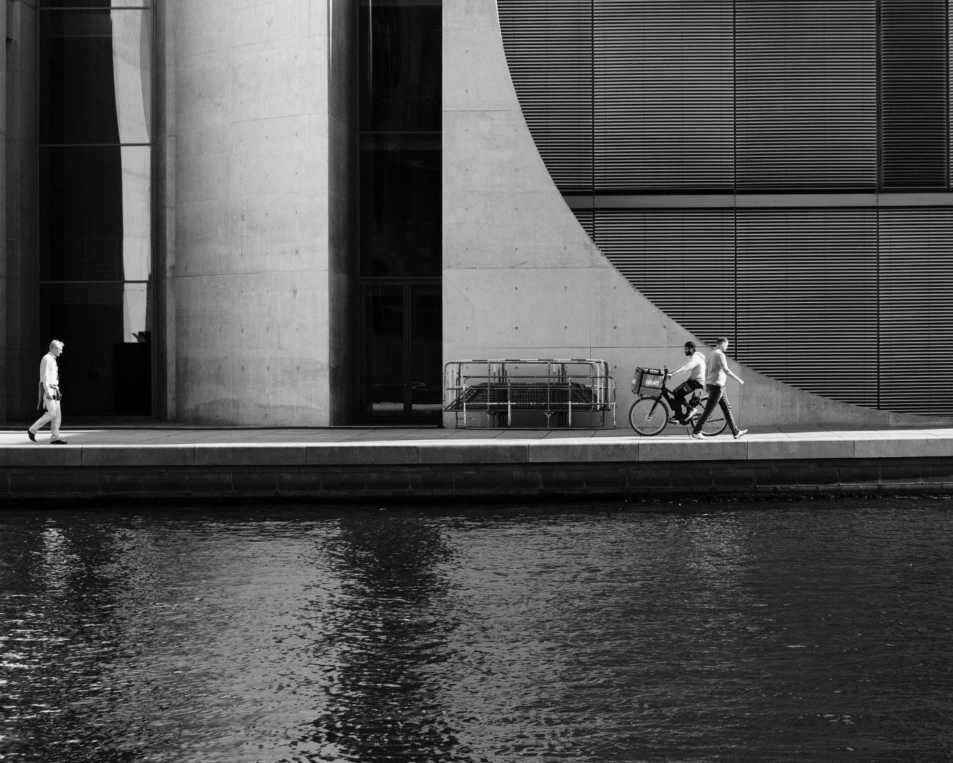 Sort-hvid foto af tre personer på fortovet ved en moderne bygning ved vandet, hvoraf en er på cykel og den tredje går, med refleksioner i vandet.