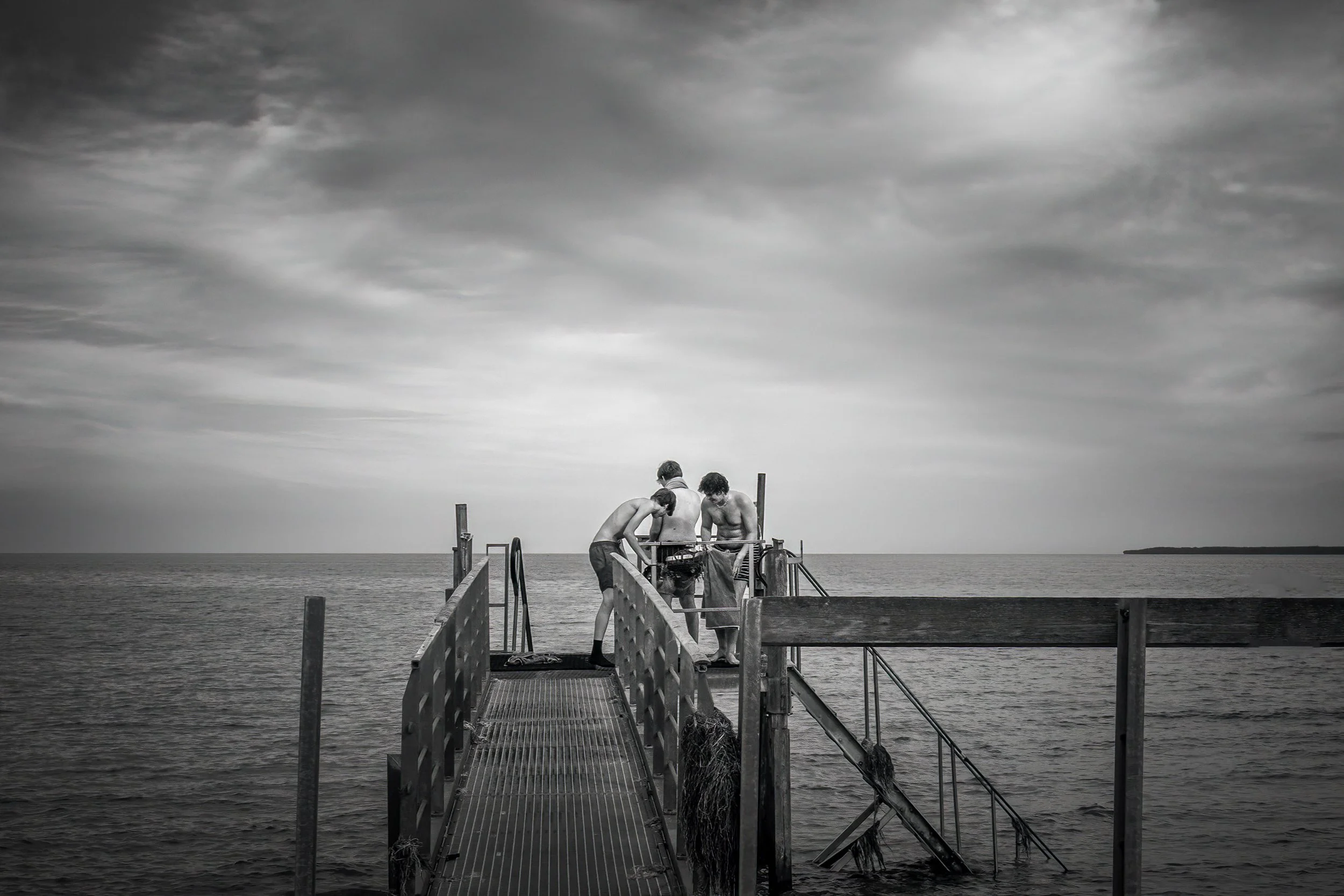 Sommer ved havet – unge mænd ved badebroen i et roligt øjeblik. Tre unge mænd står på en badebro og kigger ned i vandet under overskyet himmel.